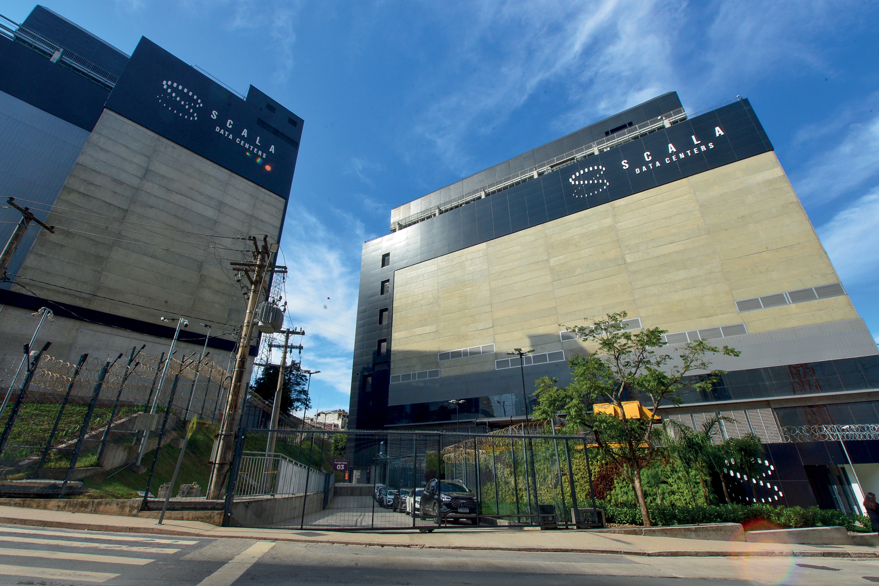 Dois prédios modernos da Scala Data Centers, com fachadas em tons de cinza e preto, sob um céu azul com nuvens esparsas. Postes de energia e fiação visíveis em primeiro plano, com uma rua e faixa de pedestres.