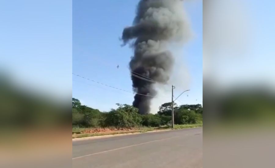 Uma densa coluna de fumaça preta sobe em espiral no céu azul claro, vista de uma estrada asfaltada. À direita, um poste de luz com fios e, ao fundo, uma faixa de vegetação verde escura. O chão ao lado da estrada é de terra batida. O céu está limpo, sem nuvens, exceto pela fumaça.
