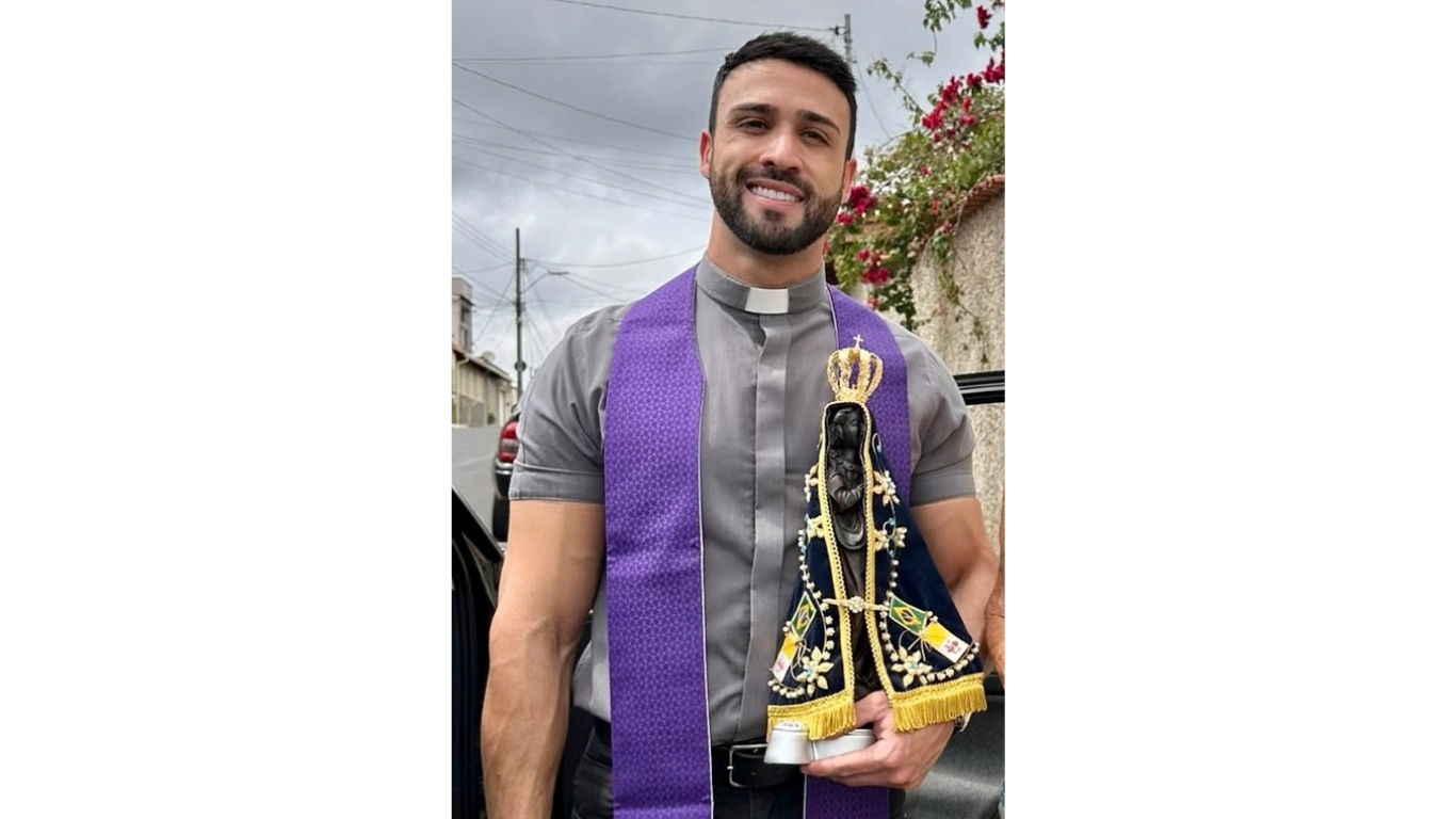 Homem jovem, barbudo, sorrindo, vestindo camisa cinza com colarinho clerical branco e estola roxa, segurando uma imagem de Nossa Senhora Aparecida com manto azul e dourado, em ambiente externo com folhagens e céu nublado