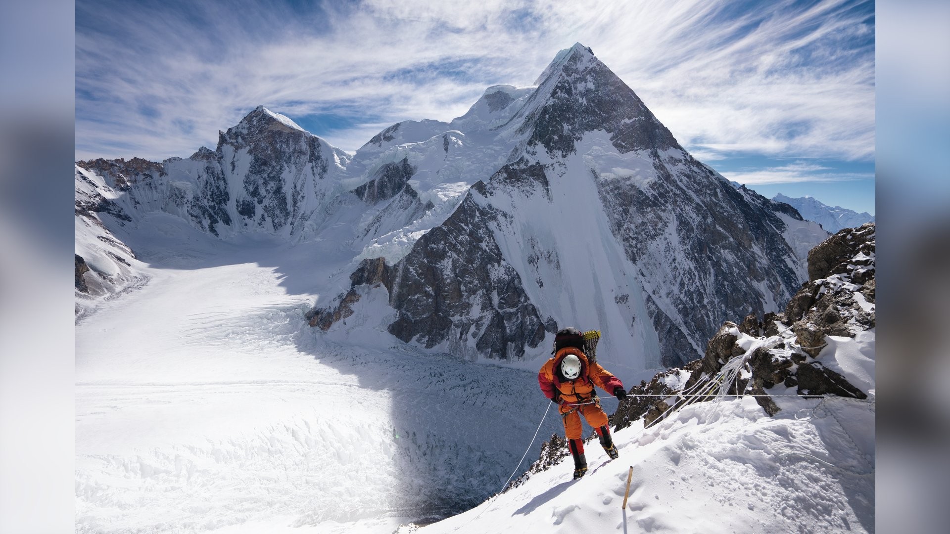 Alpinista em traje laranja e capacete branco escala uma montanha nevada, segurando uma corda. Ao fundo, picos rochosos cobertos de neve e um céu azul com nuvens esparsas.
