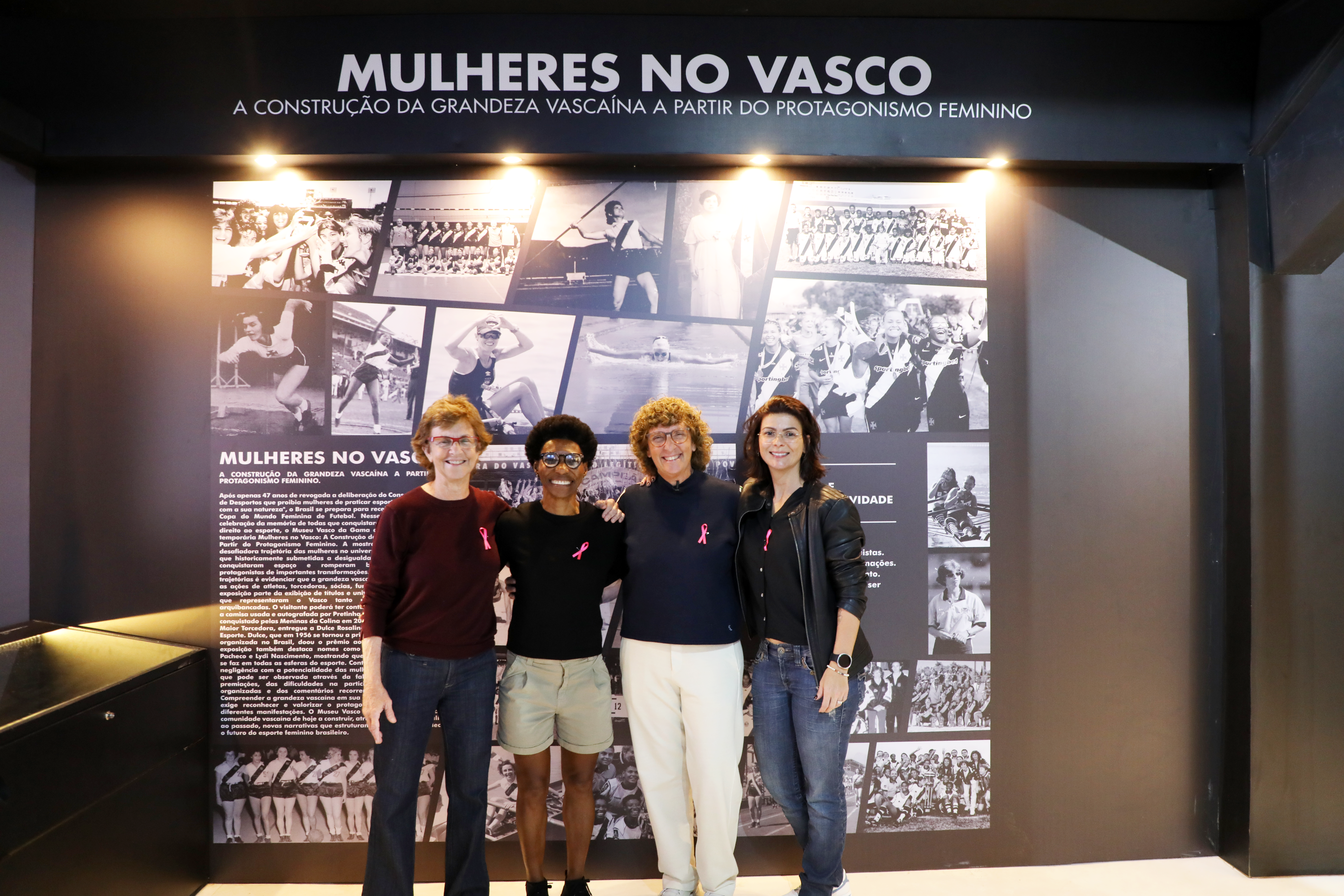 Quatro mulheres sorridentes, com laços rosa na roupa, posam em frente a um painel expositivo Mulheres no Vasco, com fotos históricas em preto e branco de atletas femininas
