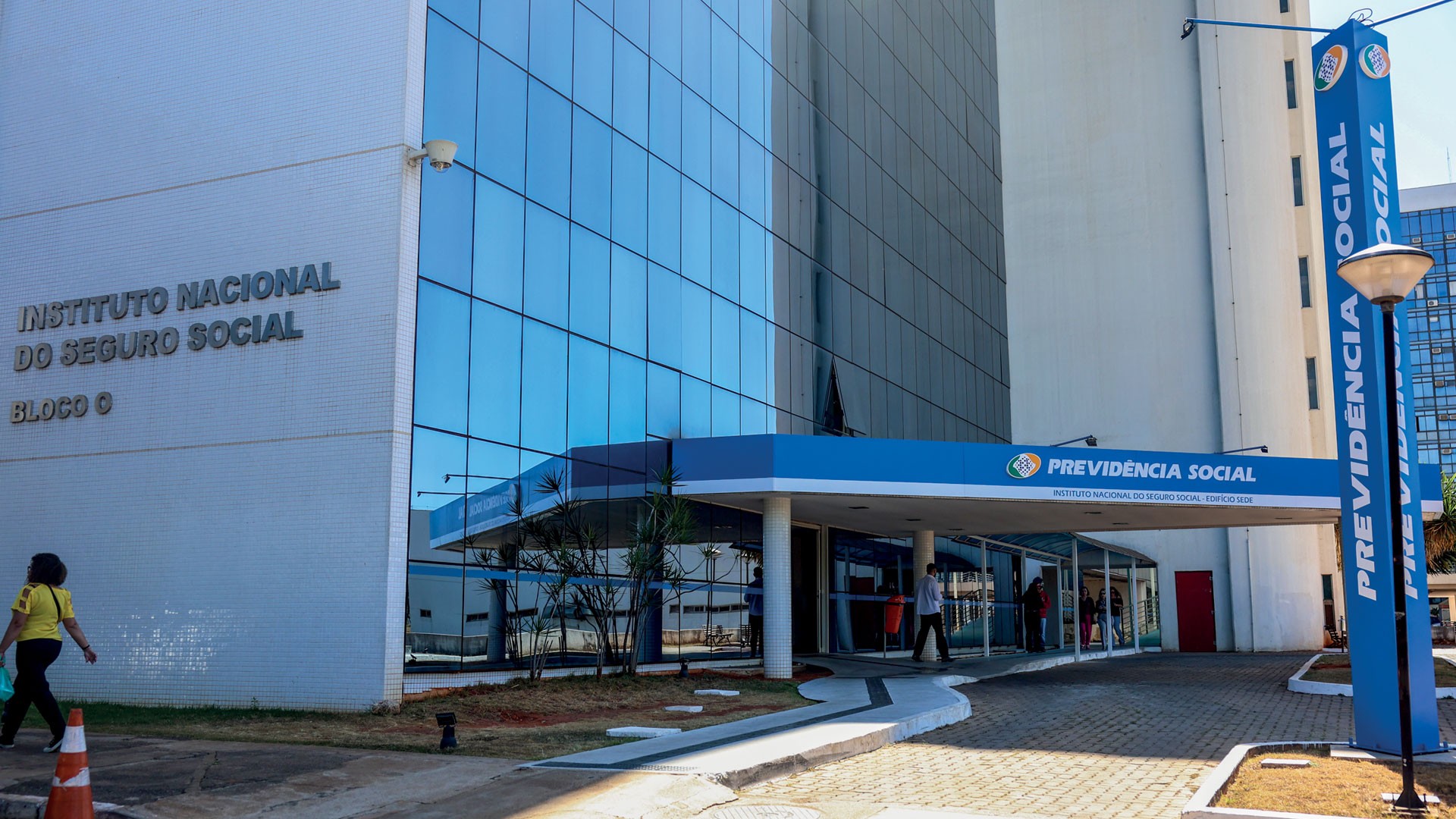 Fachada do prédio do Instituto Nacional do Seguro Social (INSS), com paredes claras e janelas espelhadas azuis. Uma mulher de camiseta amarela caminha à esquerda. Na entrada, uma placa azul indica Previdência Social.