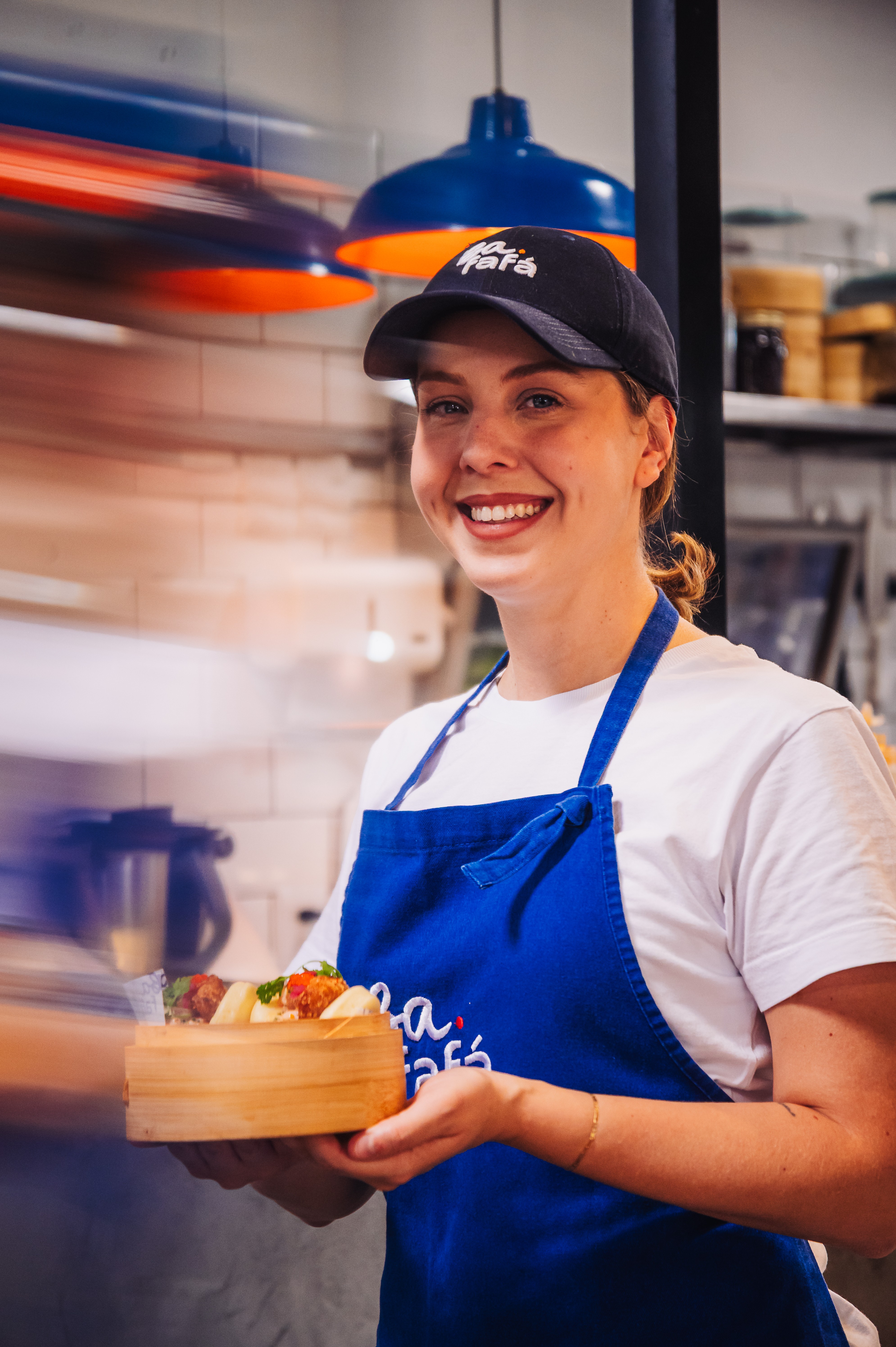 Mulher jovem sorridente, de bon&eacute; preto e avental azul com logo branco, segurando uma caixa de madeira com salgados variados
