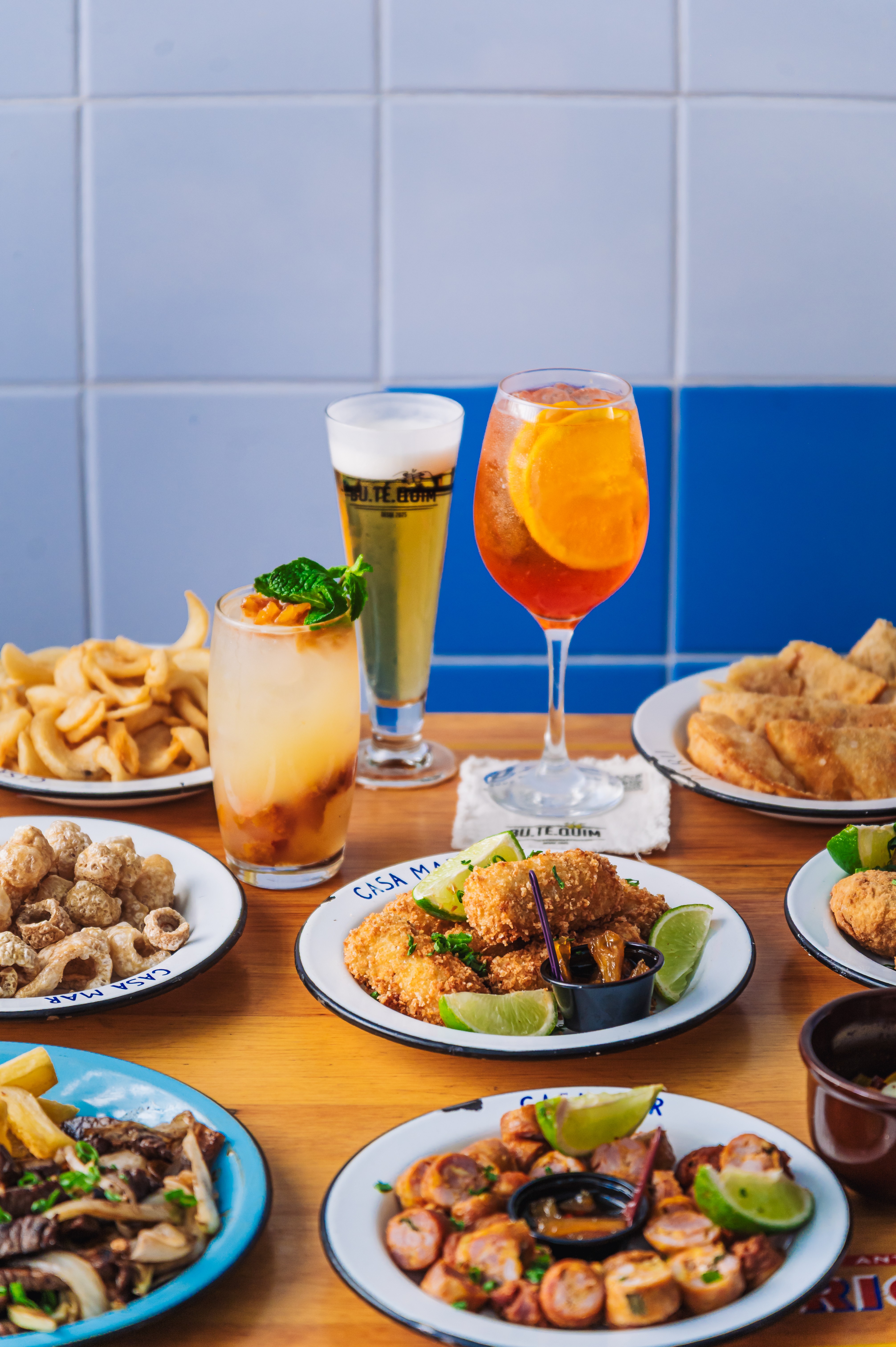 Mesa de madeira com diversos pratos de petiscos e bebidas. H&aacute; por&ccedil;&otilde;es de torresmo, batata frita, carne com cebola, lingui&ccedil;a, past&eacute;is e iscas de peixe em pratos brancos com borda preta. Tr&ecirc;s copos de bebidas: um coquetel com hortel&atilde;, uma cerveja e um Aperol Spritz com fatia de laranja. Fundo de parede com azulejos azuis e brancos
