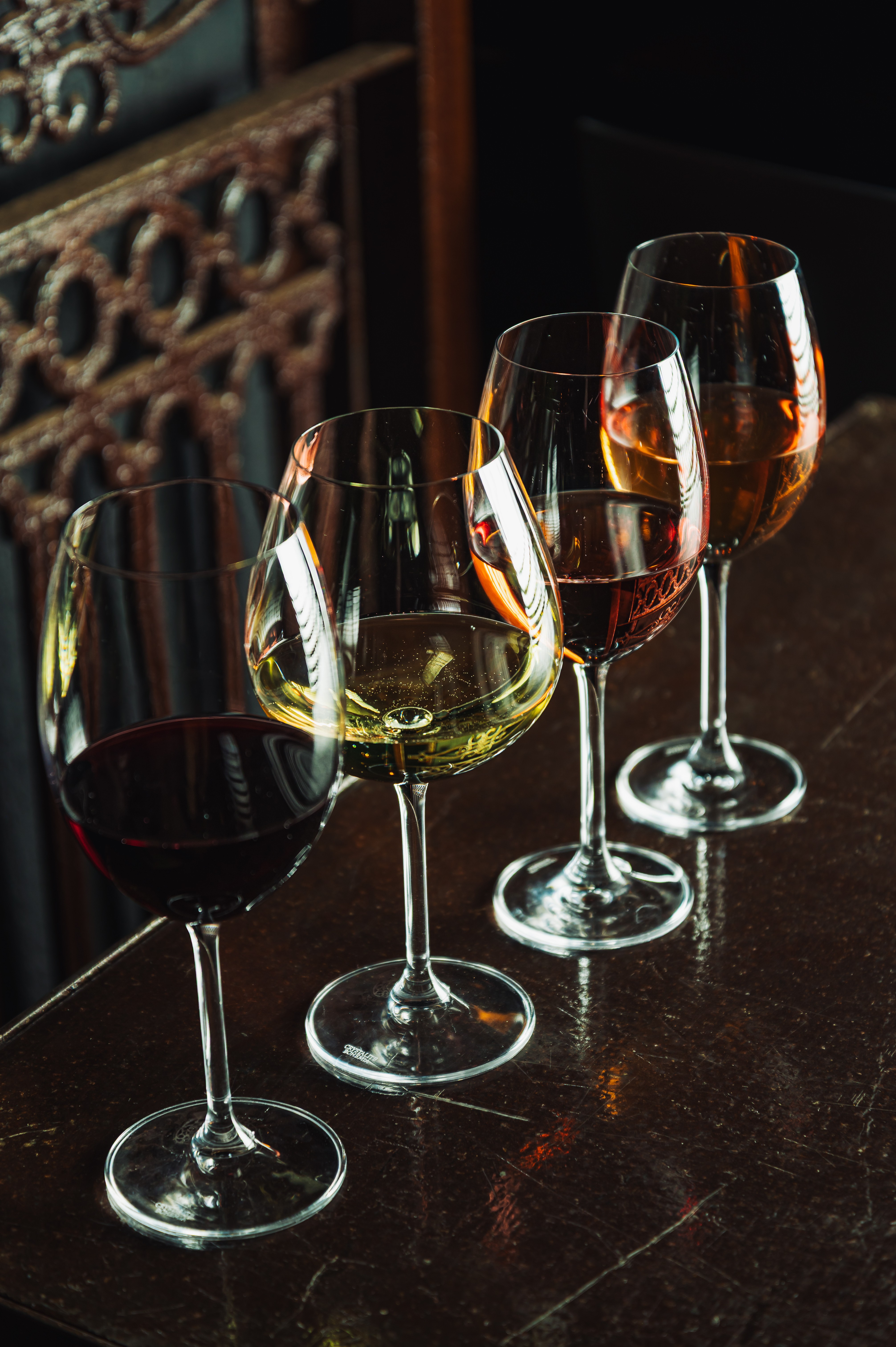 Quatro ta&ccedil;as de vinho alinhadas em uma mesa escura, da esquerda para a direita: vinho tinto, branco, ros&eacute; e &acirc;mbar, com reflexos de luz