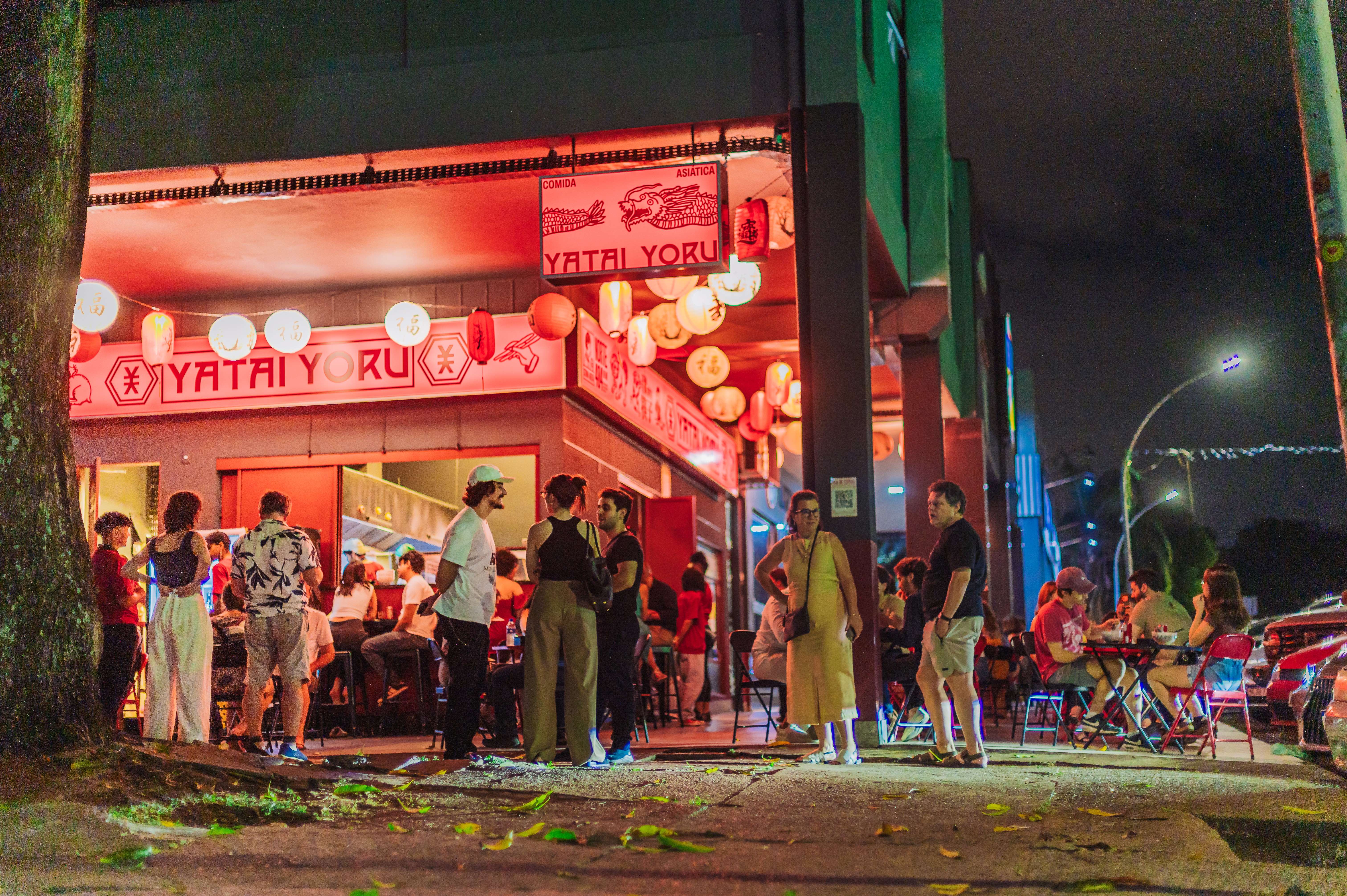 Pessoas em frente ao restaurante japon&ecirc;s Yatai Yoru, iluminado por luzes vermelhas e lanternas orientais, &agrave; noite. Algumas pessoas conversam na cal&ccedil;ada, outras sentam em mesas externas, criando um ambiente movimentado e acolhedor