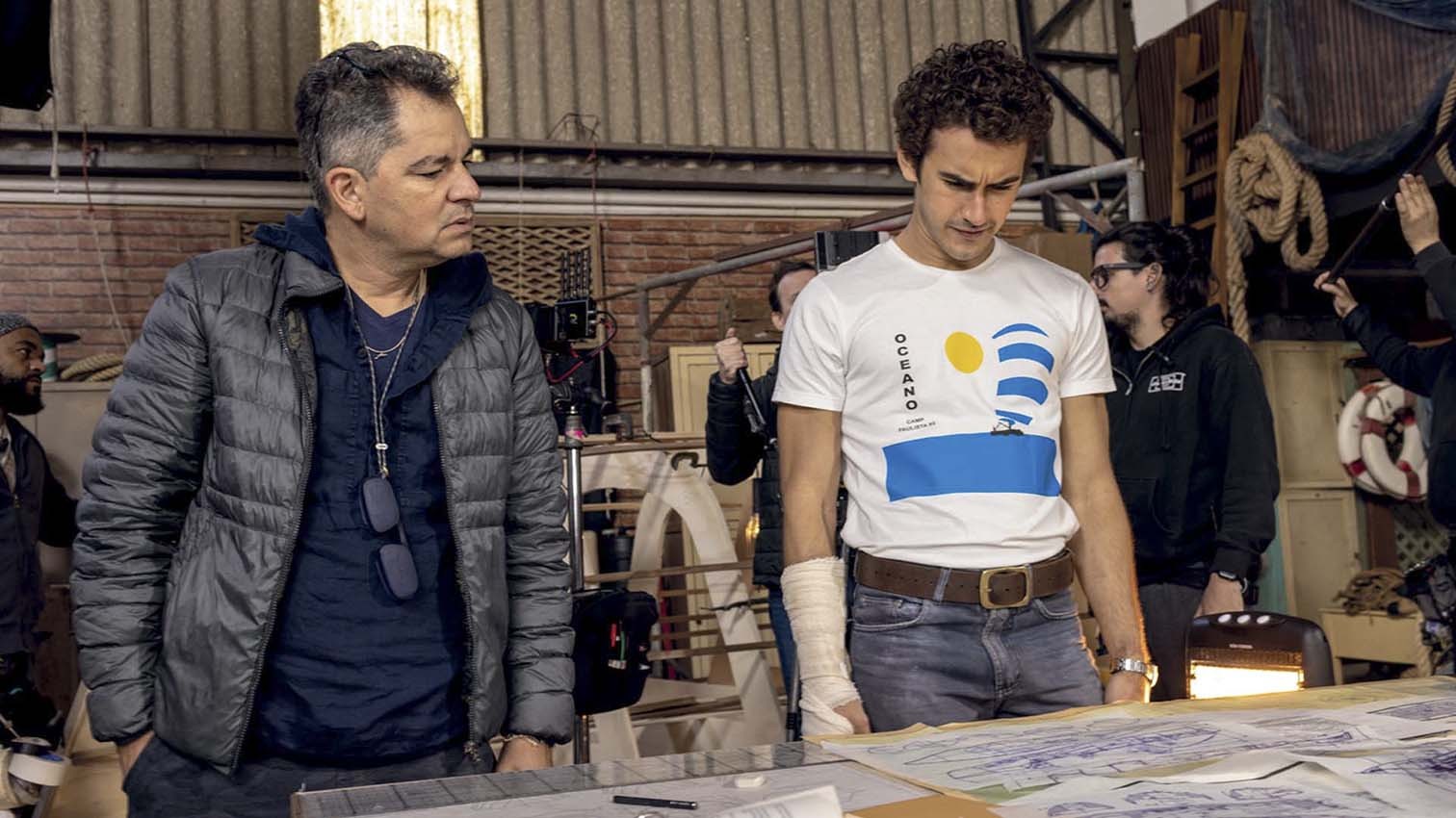 Dois homens em um set de filmagem. À esquerda, um homem de meia-idade com jaqueta cinza e óculos pendurados no pescoço. À direita, um homem jovem de camiseta branca com estampa de barco e faixa azul, e jeans, com o braço enfaixado. Ambos olham para baixo, concentrados em papéis sobre uma mesa. Ao fundo, equipamentos de filmagem e outros membros da equipe