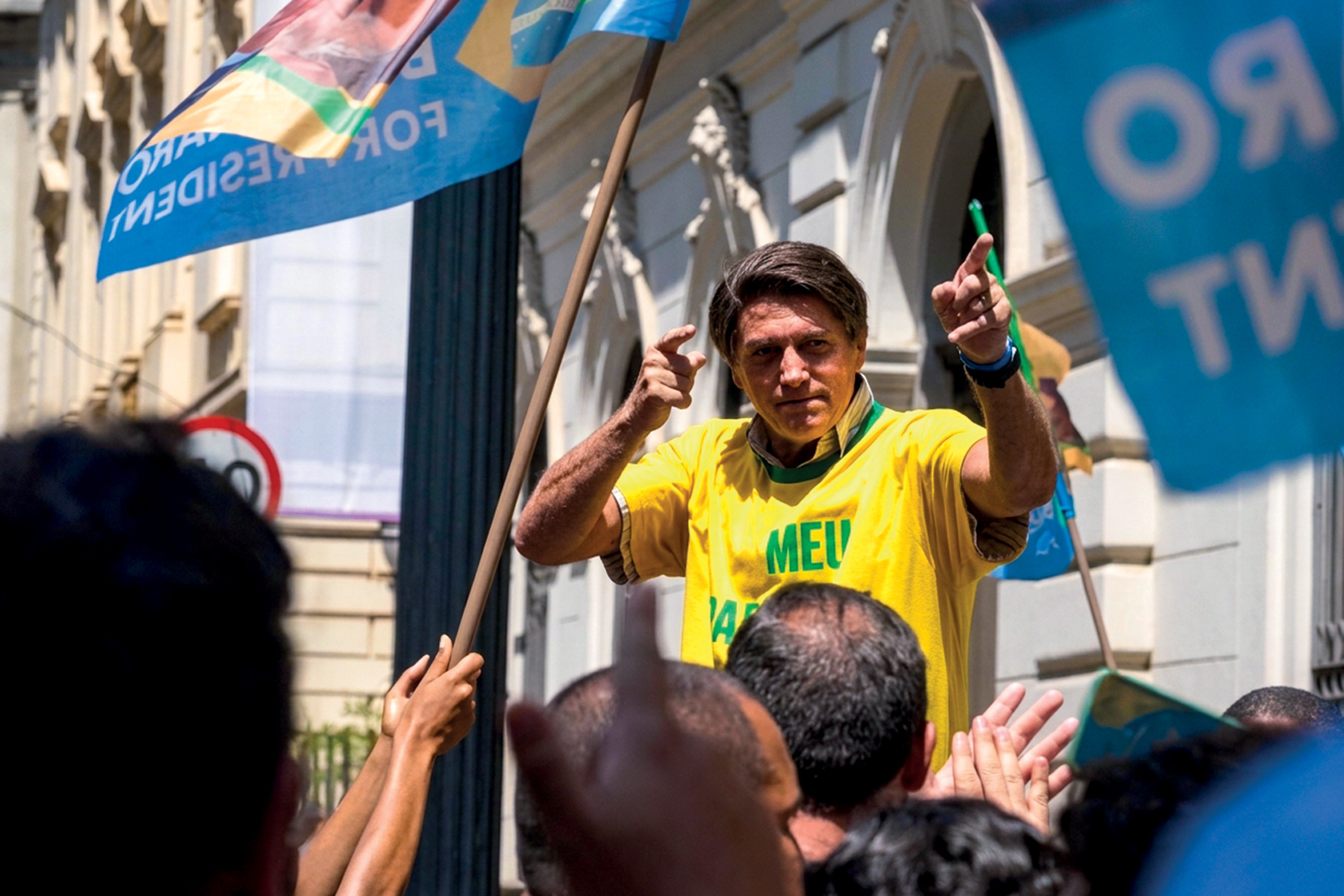 Jair Bolsonaro, vestindo camisa amarela da seleção brasileira, gesticula com as mãos para uma multidão, com bandeiras azuis de campanha ao redor, em um evento político diurno