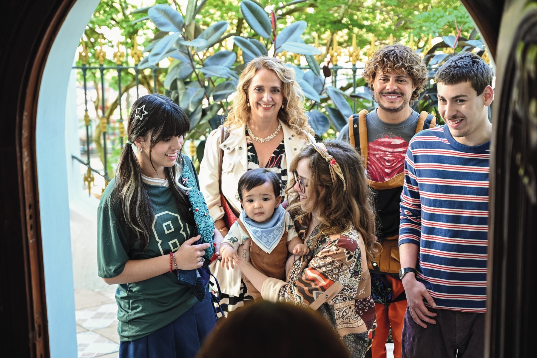 Cinco adultos e um bebê sorrindo, em pé, olhando para a frente, em um arco de porta. A mulher loira no centro usa um colar e casaco claro. O bebê no colo de uma mulher de óculos veste macacão marrom e babador azul. Dois homens estão atrás, um com camiseta estampada e outro com listras azuis e vermelhas. Uma jovem com maria-chiquinha e camiseta verde está à esquerda. Folhagens verdes ao fundo