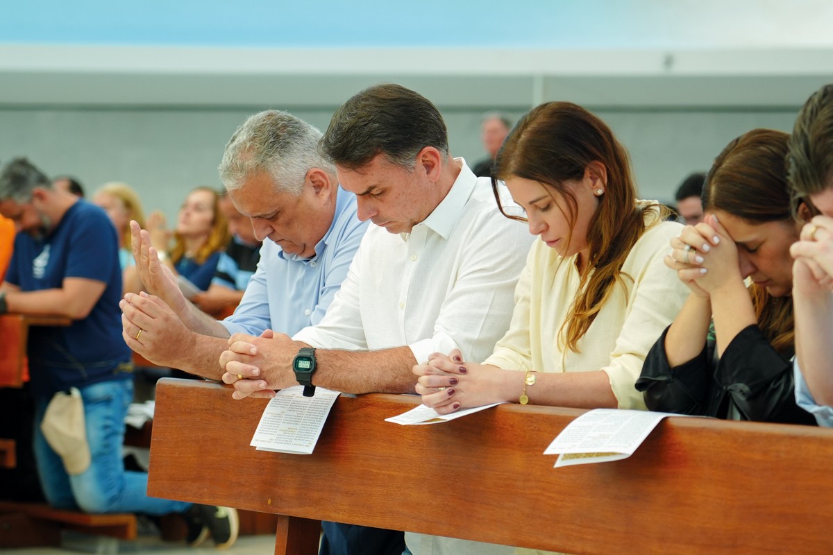 Pessoas em um banco de igreja, com cabeças baixas e mãos unidas em oração. Um homem de camisa branca e outro de camisa azul clara estão no centro, ao lado de duas mulheres, uma de blusa creme e outra de blusa escura. Folhetos brancos estão sobre o banco de madeira.