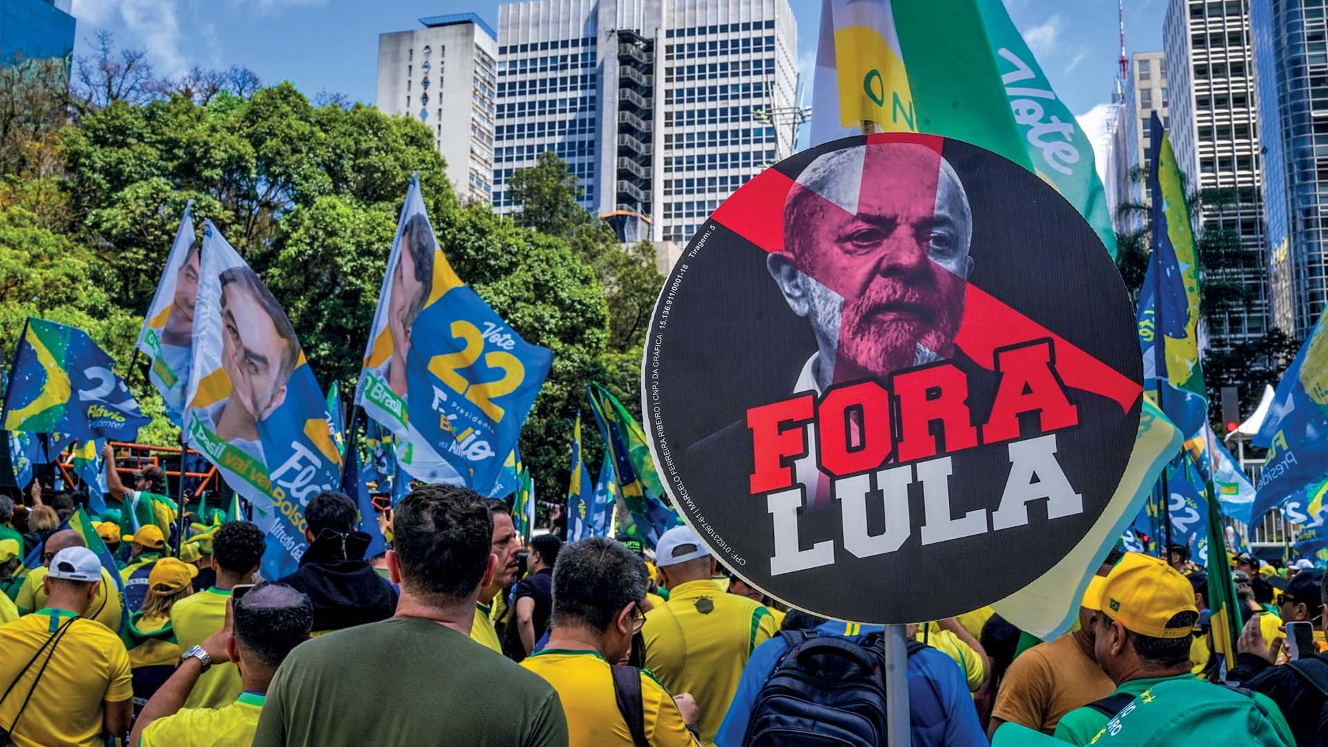 Manifestantes em um protesto político, com um cartaz grande e redondo em primeiro plano exibindo a imagem de um homem barbudo em tons de vermelho e branco, com o texto FORA LULA em letras maiúsculas vermelhas e brancas. Ao fundo, bandeiras azuis e amarelas com o número 22 e rostos de políticos, além de bandeiras do Brasil, árvores e edifícios altos.