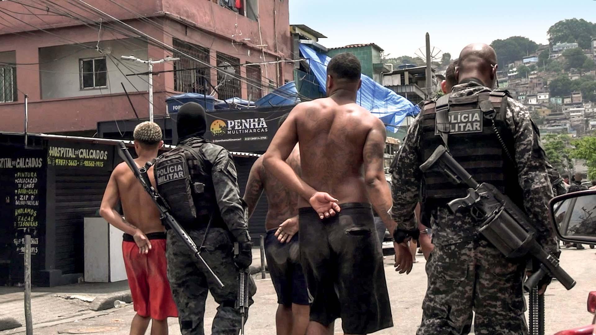 Policiais militares armados com fuzis escoltam homens sem camisa e com as mãos para trás em uma rua de comunidade, com casas e comércios ao fundo