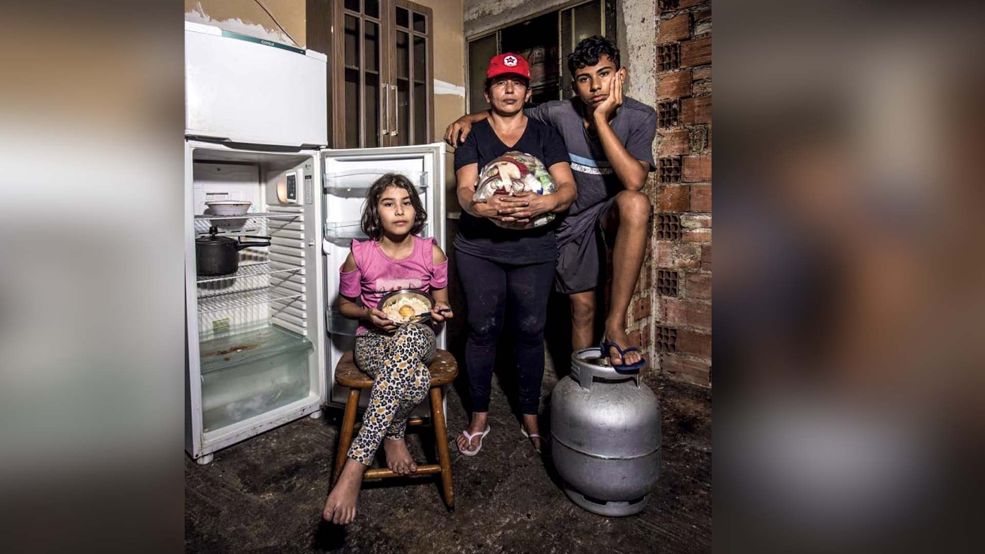 Família de três pessoas em casa simples, com geladeira aberta e vazia. Uma mulher de boné vermelho e blusa preta segura uma sacola de lixo reciclável. Uma menina sentada em um banco, com blusa rosa e calça estampada, segura um prato com um ovo frito. Um adolescente de camiseta cinza e bermuda escura apoia o pé em um botijão de gás. O chão está molhado e o fundo tem parede de tijolos.