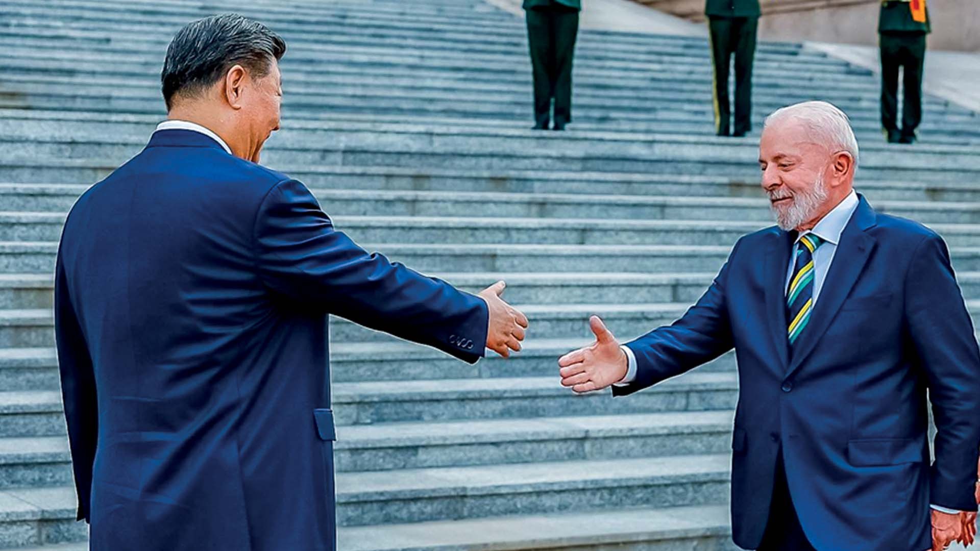 O presidente chinês Xi Jinping, de costas, e o presidente brasileiro Lula, de perfil, estendem as mãos para um aperto, ambos vestindo ternos azuis escuros. Eles estão em frente a uma escadaria de pedra, com duas pessoas em uniforme militar ao fundo