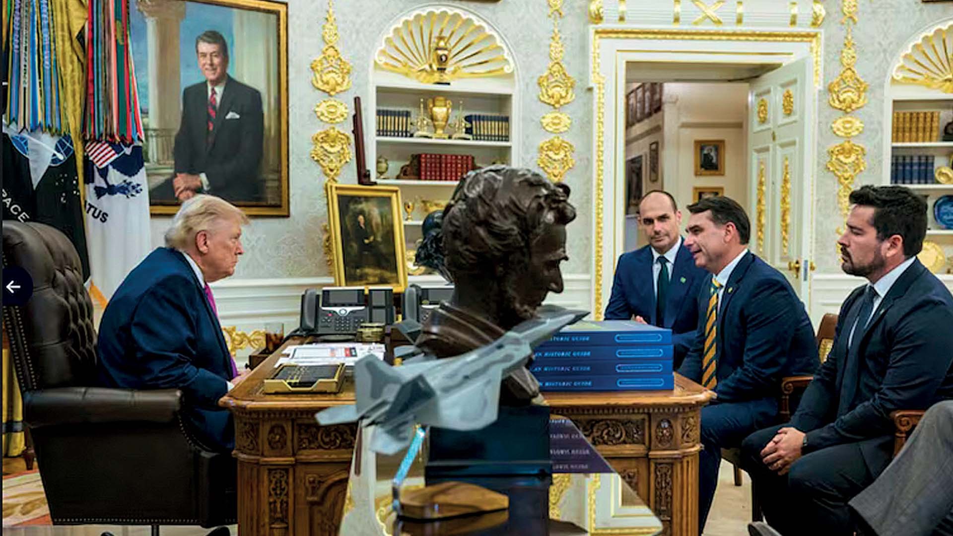 Donald Trump, de costas, sentado em uma poltrona de couro, conversa com três homens de terno sentados à sua frente em um escritório luxuoso. Sobre a mesa, uma escultura de busto e um avião em miniatura. Ao fundo, um quadro de Ronald Reagan.