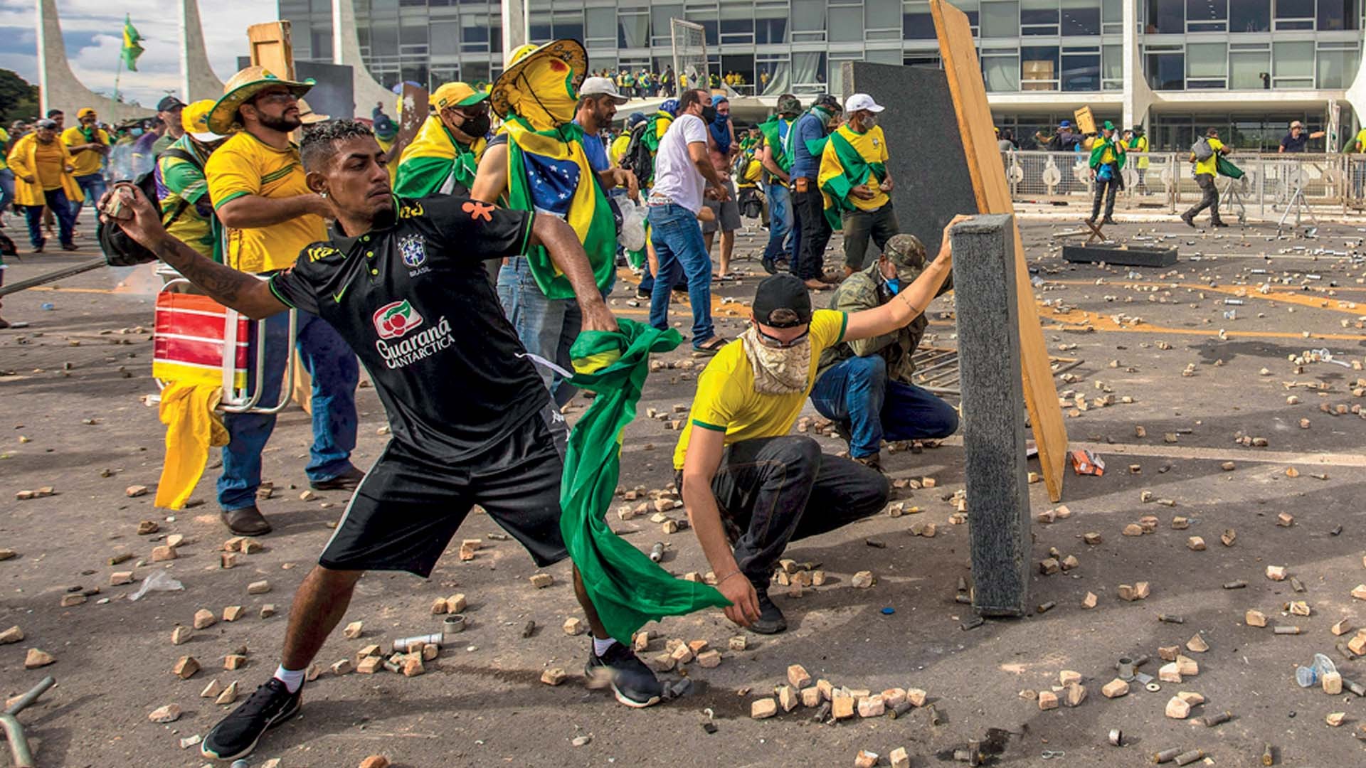 Manifestantes em frente a um prédio governamental, com um homem de camisa preta arremessando um objeto e outro agachado, pegando pedaços de madeira no chão. O asfalto está coberto por detritos e pedras, e outros manifestantes com roupas verde-amarelas estão ao fundo, alguns com o rosto coberto por bandeiras.