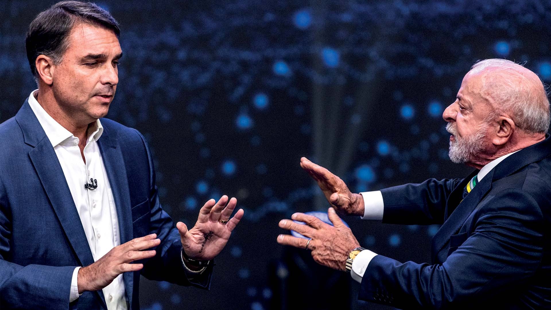 Dois homens, um de terno azul e camisa branca, e outro de terno azul com gravata verde e amarela, gesticulam com as mãos abertas em um debate, contra um fundo escuro com pontos de luz azuis