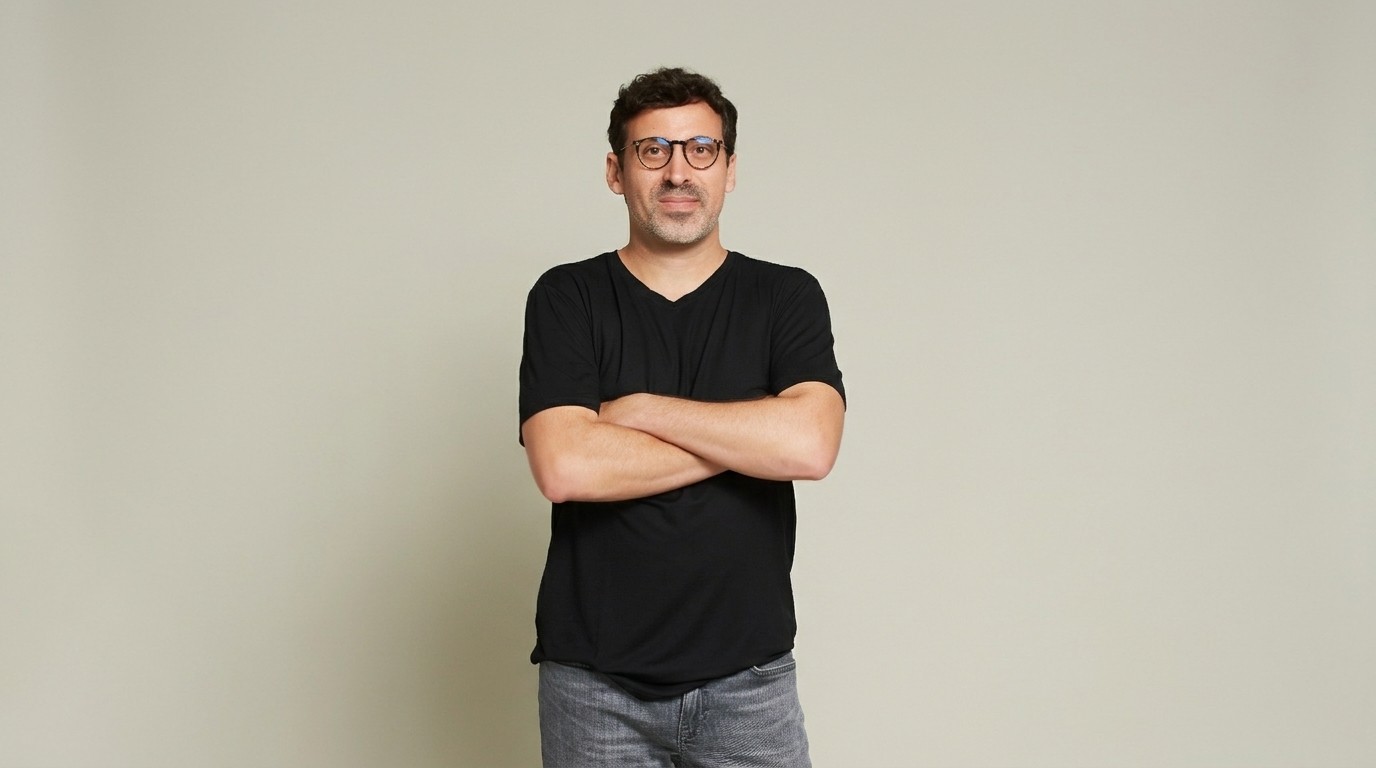 Homem de pele clara, cabelo escuro e óculos redondos, vestindo camiseta preta e calça jeans cinza, braços cruzados, olhando para frente com expressão neutra, em fundo bege claro