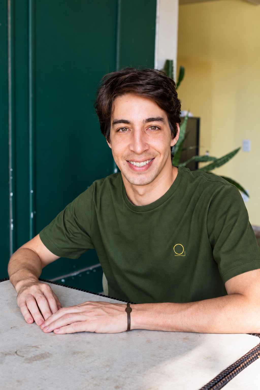 Homem jovem de pele clara, cabelo castanho escuro e sorriso largo, vestindo camiseta verde-oliva com um c&iacute;rculo amarelo no peito, sentado &agrave; mesa com as m&atilde;os entrela&ccedil;adas. Ao fundo, uma porta verde escura e uma parede amarela com uma planta verde