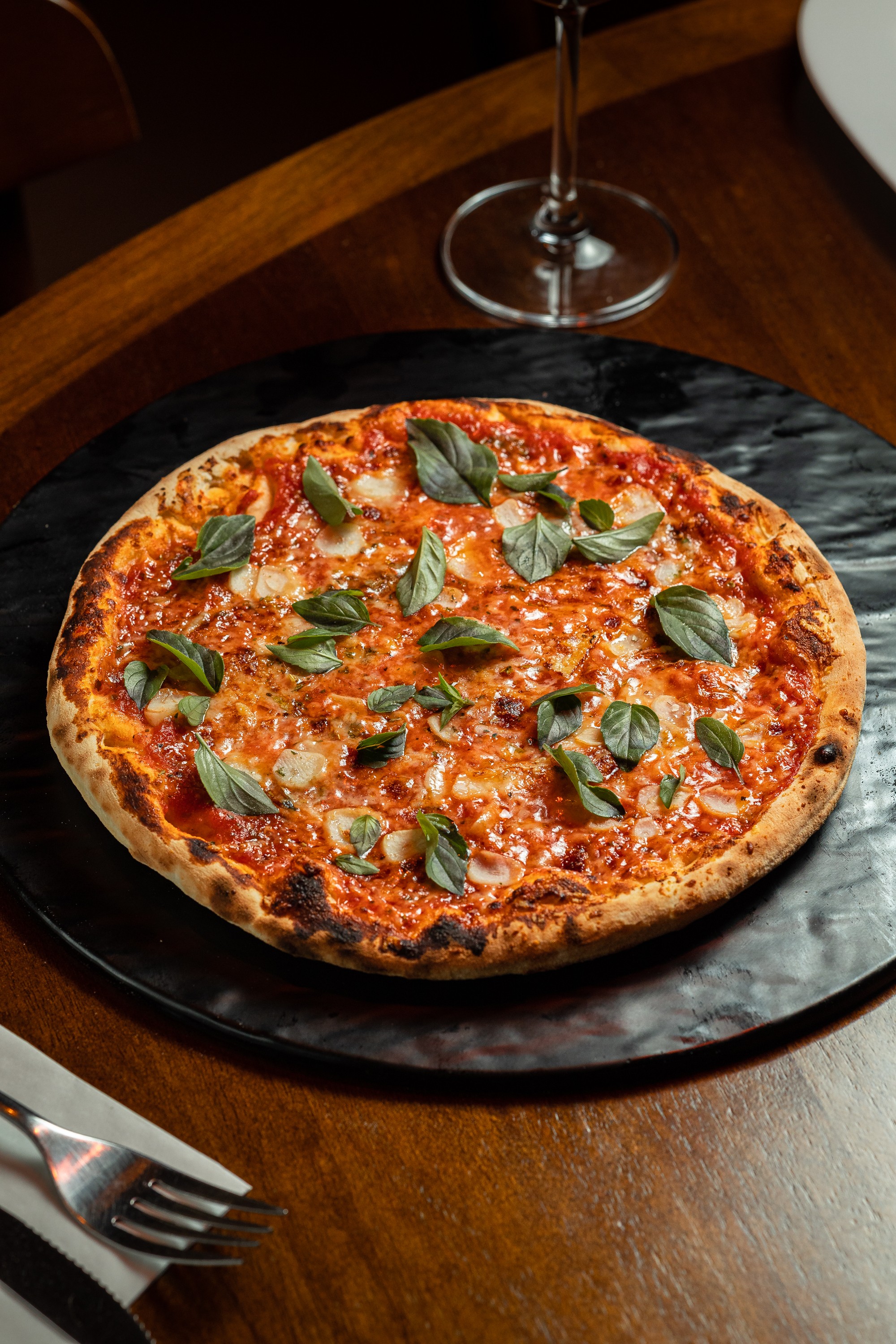 Pizza margherita redonda com borda crocante, molho de tomate, queijo derretido e folhas de manjeric&atilde;o fresco, servida em prato preto sobre mesa de madeira escura com talheres e ta&ccedil;a de vinho ao fundo
