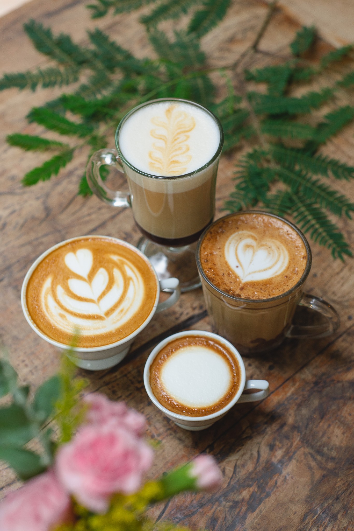 Quatro x&iacute;caras de caf&eacute; com leite e arte latte sobre uma mesa de madeira r&uacute;stica, com ramos verdes e flores cor-de-rosa desfocadas em primeiro plano. A x&iacute;cara superior tem um desenho de folha, a do meio &agrave; esquerda um cora&ccedil;&atilde;o com folhas, a do meio &agrave; direita um cora&ccedil;&atilde;o simples, e a inferior um c&iacute;rculo de espuma.