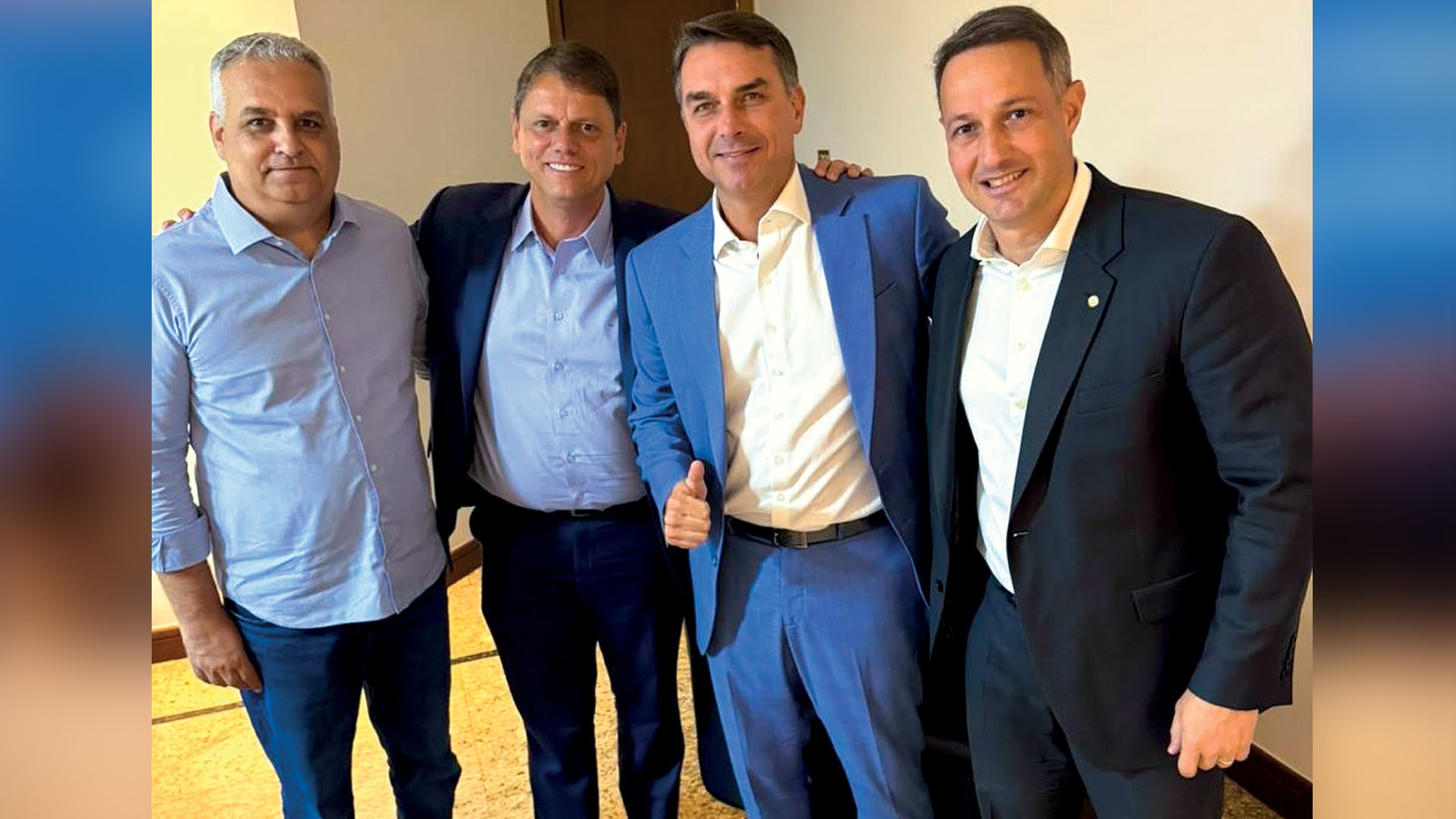 Quatro homens sorrindo, em pé, posam para foto. O segundo da direita, de terno azul, faz um sinal de positivo com o polegar