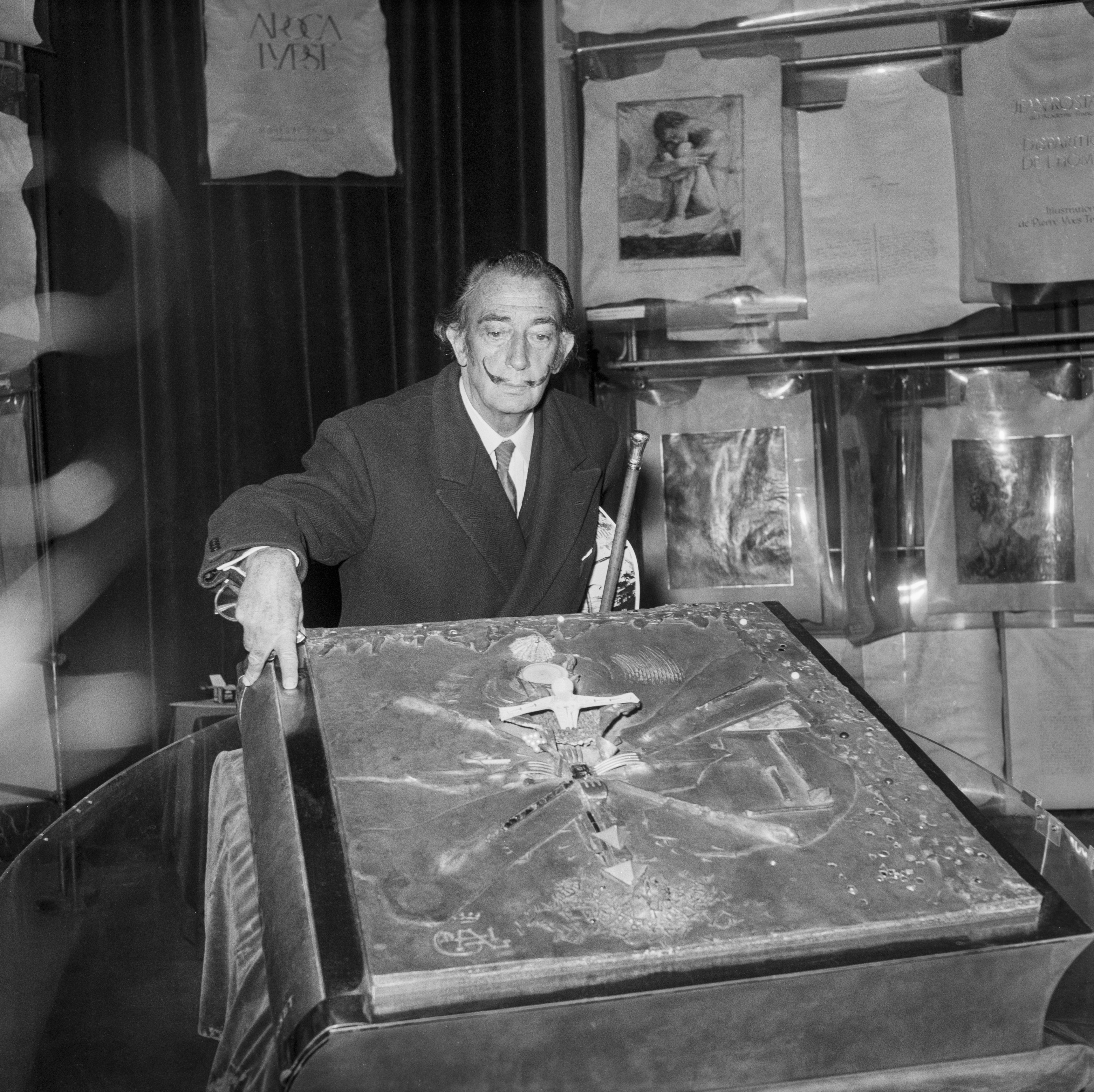 Salvador Dalí, com seu bigode icônico, inclina-se sobre um livro grande e ornamentado, com uma cruz em relevo na capa. Ele veste um terno escuro e gravata, segurando uma bengala. Ao fundo, sacolas de tecido com a inscrição ARCA IVEST e quadros emoldurados estão expostos