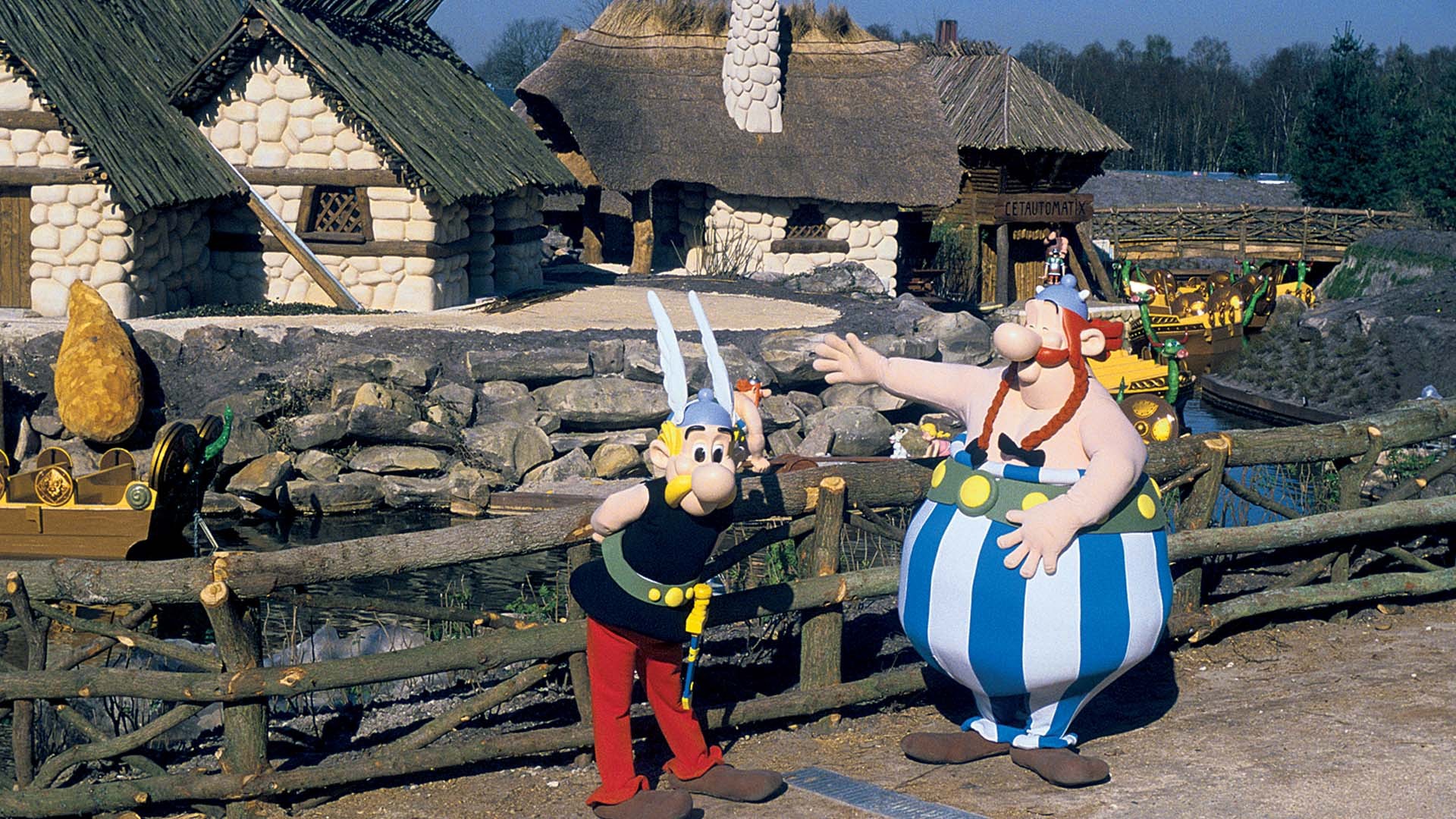Estátuas de Astérix e Obélix em um parque temático, com Obélix gesticulando e Astérix ao seu lado. Ao fundo, casas rústicas de pedra com telhados de palha e um barco no lago, simulando uma vila gaulesa.
