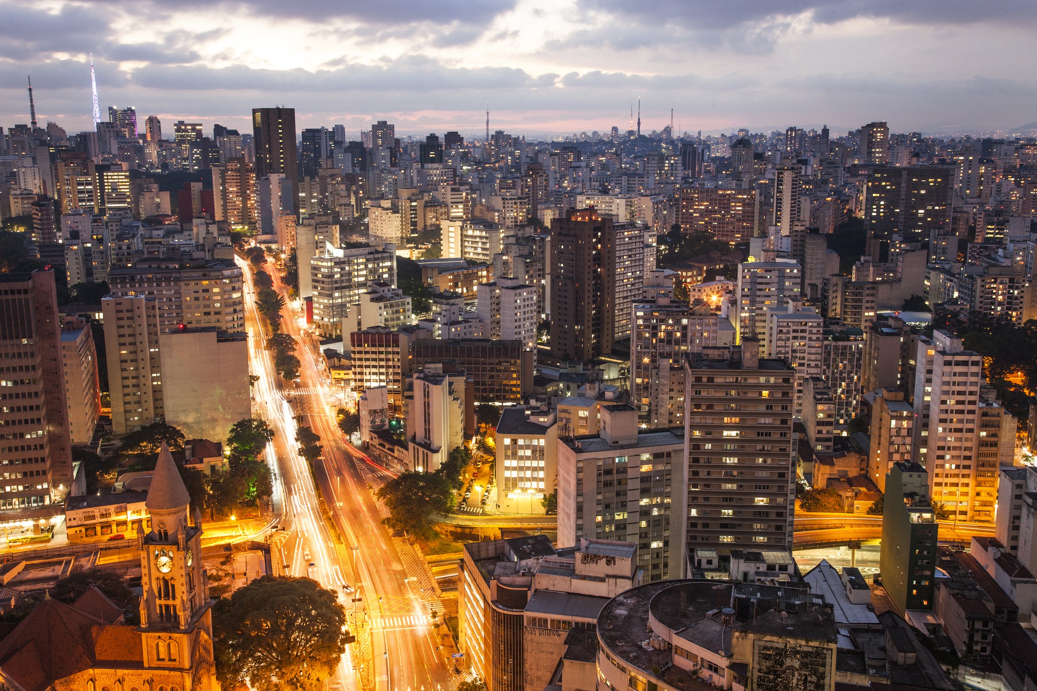 Vista aérea noturna de uma cidade grande, com arranha-céus iluminados e uma avenida central com rastros de luzes de veículos, sob um céu nublado ao anoitecer