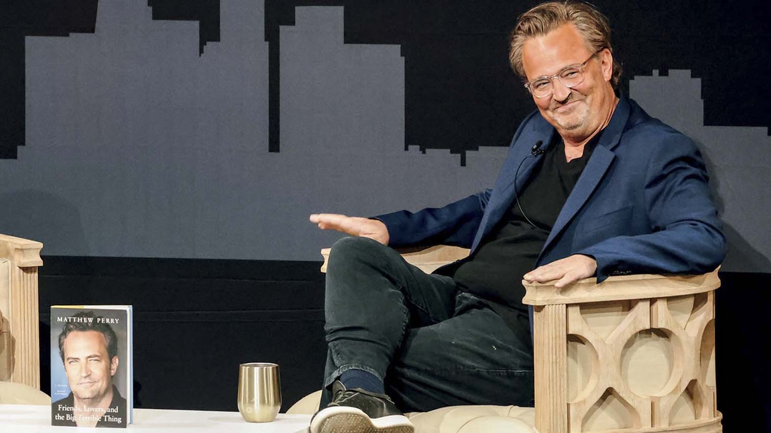 Matthew Perry, sorridente e de óculos, sentado em uma poltrona clara, vestindo blazer azul e camiseta preta. Ao lado, seu livro Friends, Lovers, and the Big Terrible Thing e um copo dourado. O fundo é cinza escuro com silhuetas de prédios