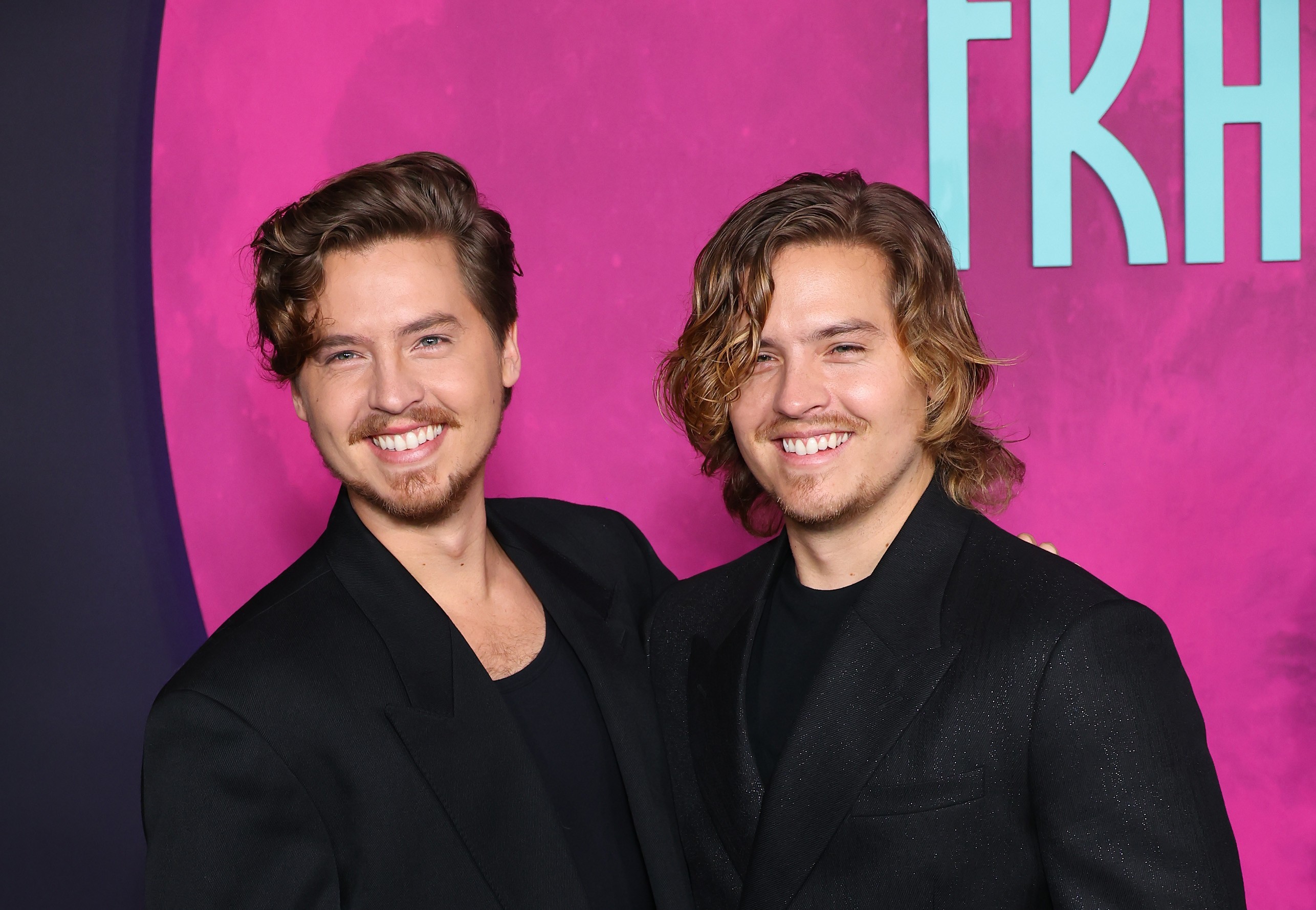 Os irmãos gêmeos Dylan e Cole Sprouse sorriem para a câmera, usando blazers pretos sobre camisetas escuras, em um fundo rosa com letras azuis