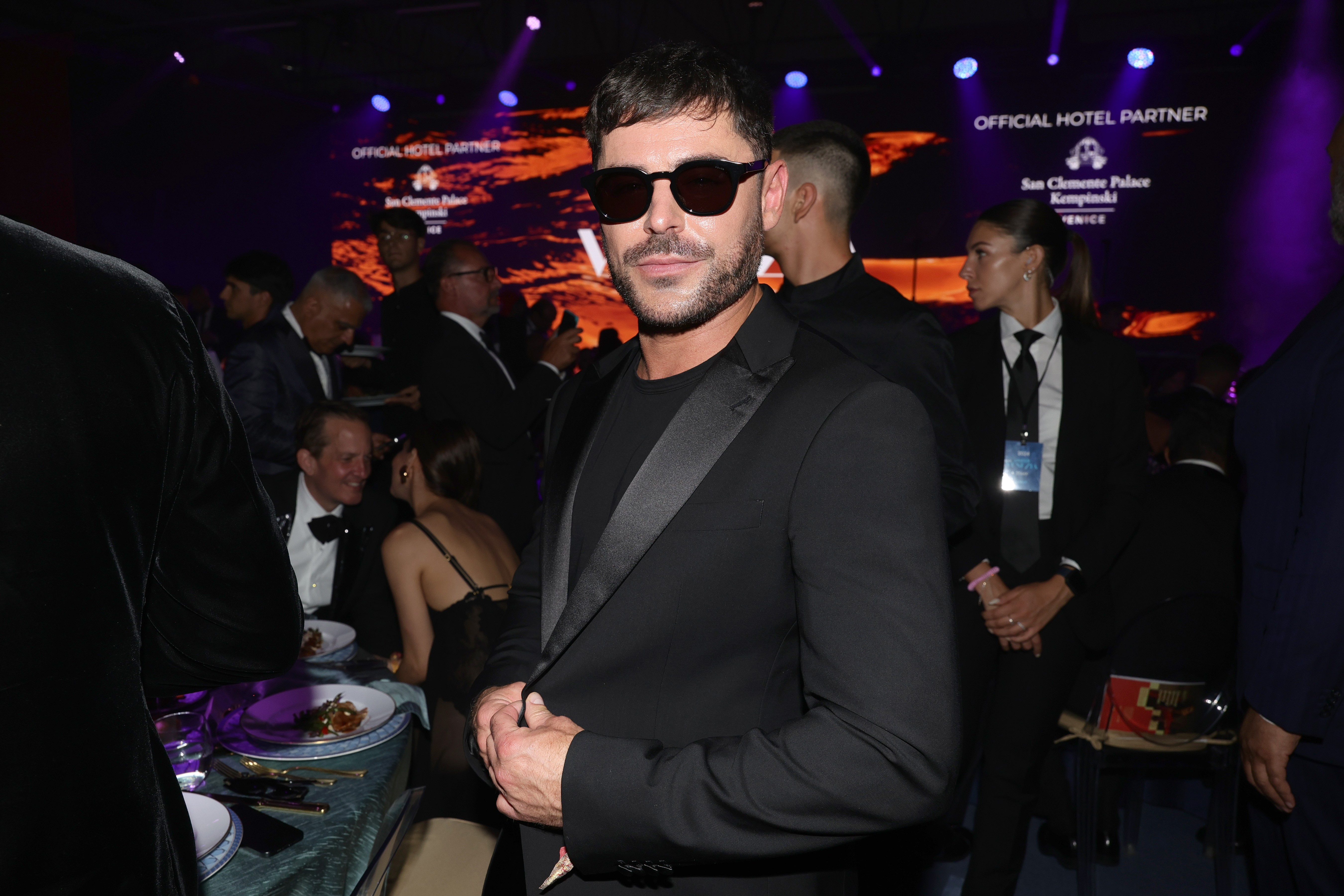 Zac Efron, com barba e óculos escuros, veste terno preto e camiseta preta, posando em evento noturno. Ao fundo, pessoas sentadas em mesas e um telão com a frase OFFICIAL HOTEL PARTNER e o nome de um hotel