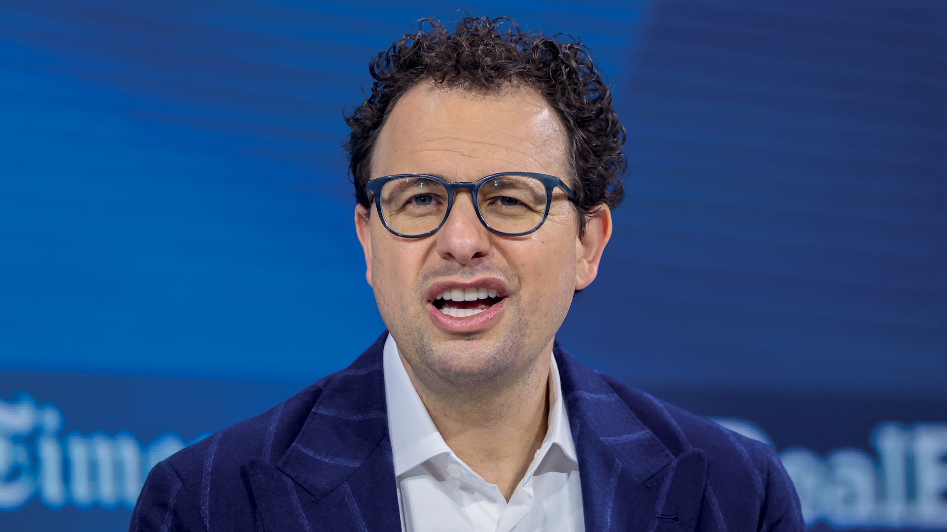 Homem de pele clara, cabelo cacheado escuro e óculos azuis, vestindo blazer azul-marinho listrado e camisa branca, falando com a boca aberta contra um fundo azul com texto desfocado