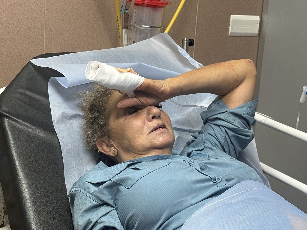 Mulher de meia-idade, cabelos cacheados e loiros, deitada em maca hospitalar com lençol azul, usando camisa azul-clara. Ela tem um curativo branco na mão direita, que está sobre a testa, cobrindo os olhos. Seu rosto expressa dor ou desconforto, com a boca entreaberta. Ao fundo, equipamentos médicos e parede bege