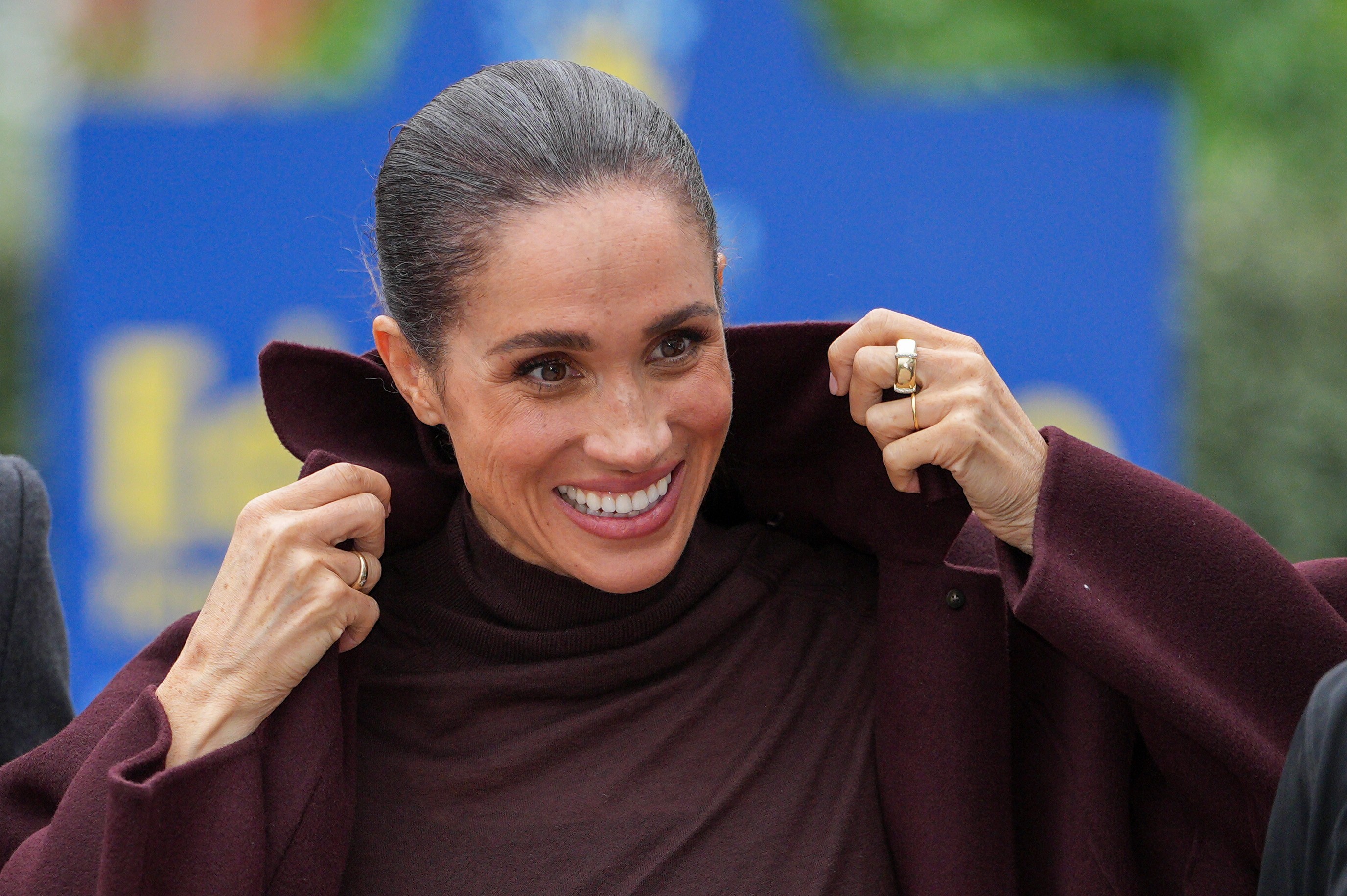 Meghan Markle sorrindo, com cabelo preso e maquiagem leve, vestindo um casaco vinho e blusa de gola alta, segurando a gola do casaco com as mãos. Ela usa anéis dourados nos dedos anelares.