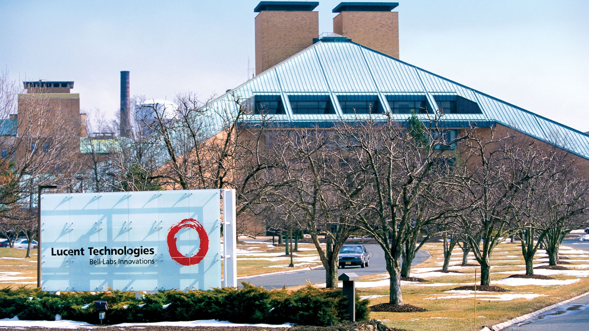 Placa branca com Lucent Technologies Bell Labs Innovations e logo circular vermelho, em frente a um prédio de tijolos com telhado inclinado e árvores secas, com neve no chão