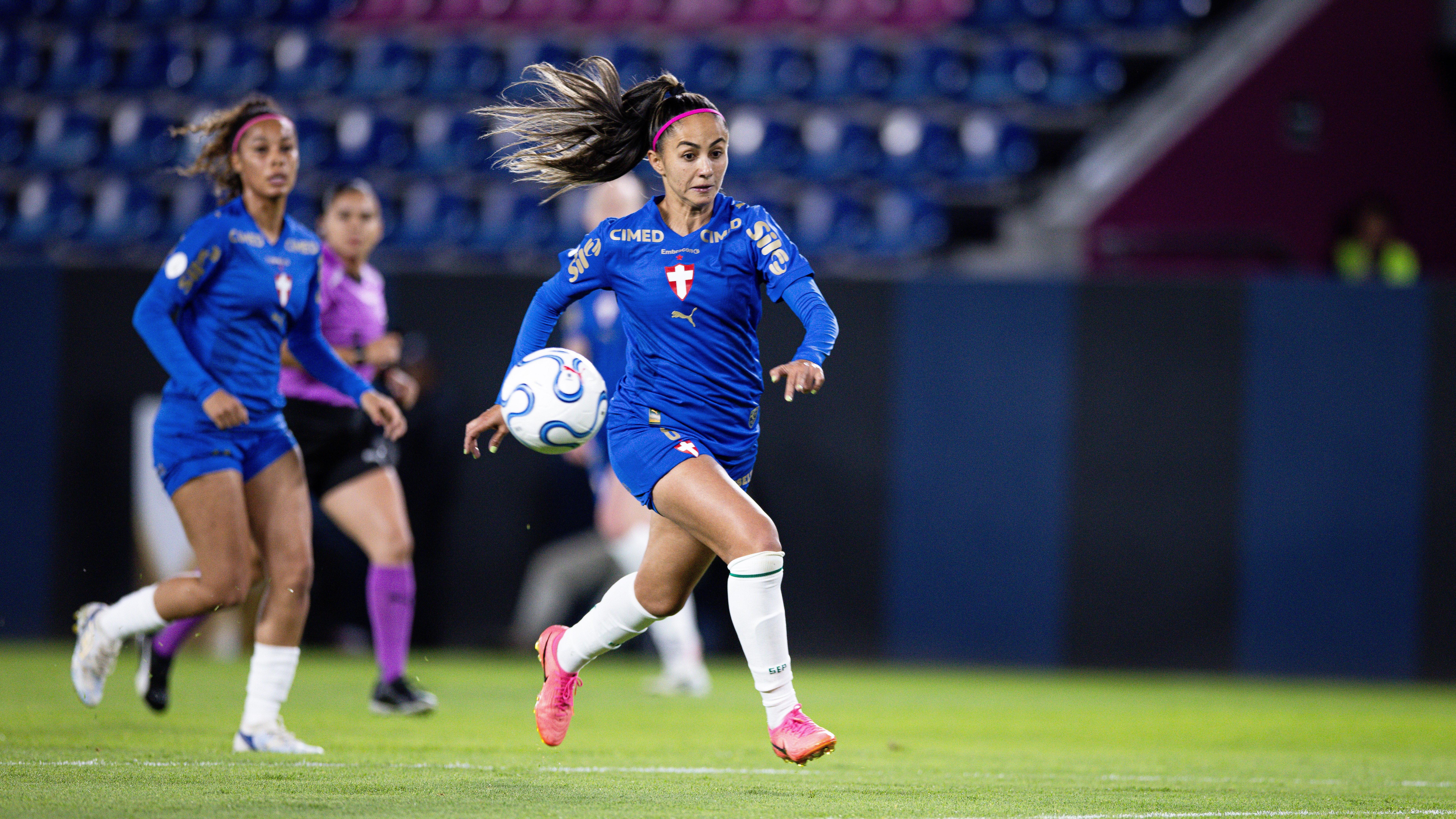 Jogadora de futebol com uniforme azul e faixa rosa no cabelo, dribla a bola com o pé direito em campo verde. Outras jogadoras desfocadas ao fundo.