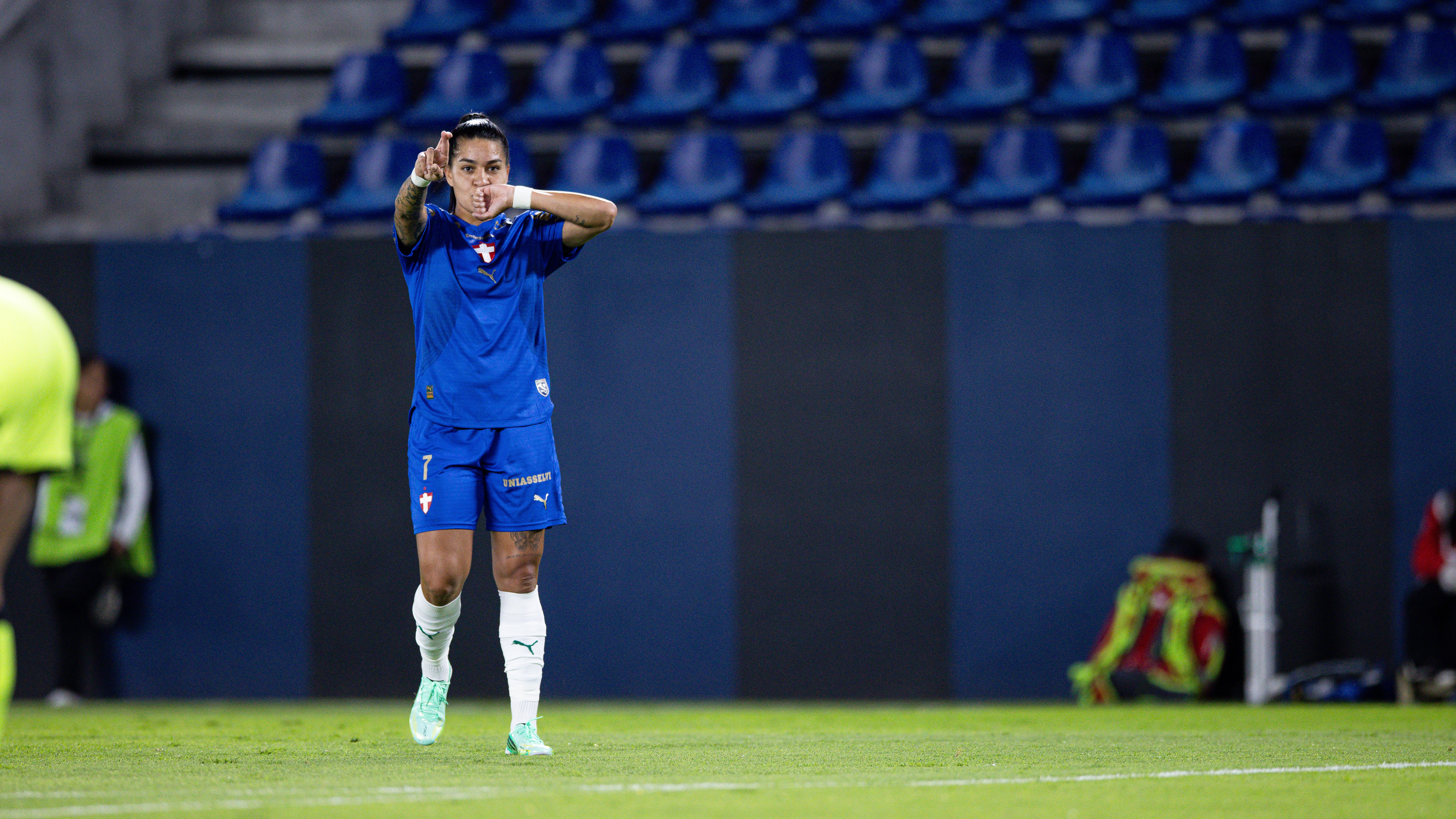 Jogadora de futebol com uniforme azul, cabelo preso, fazendo um gesto de coração com as mãos no campo, com arquibancada azul ao fundo