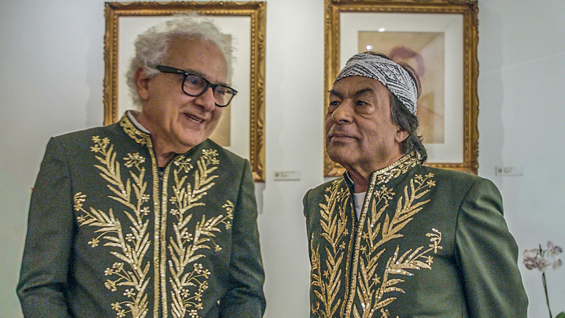 Dois homens, um de óculos e cabelo branco sorrindo, e outro de bandana e cabelo escuro, vestem casacos verde-escuros com bordados dourados, em frente a uma parede branca com quadros