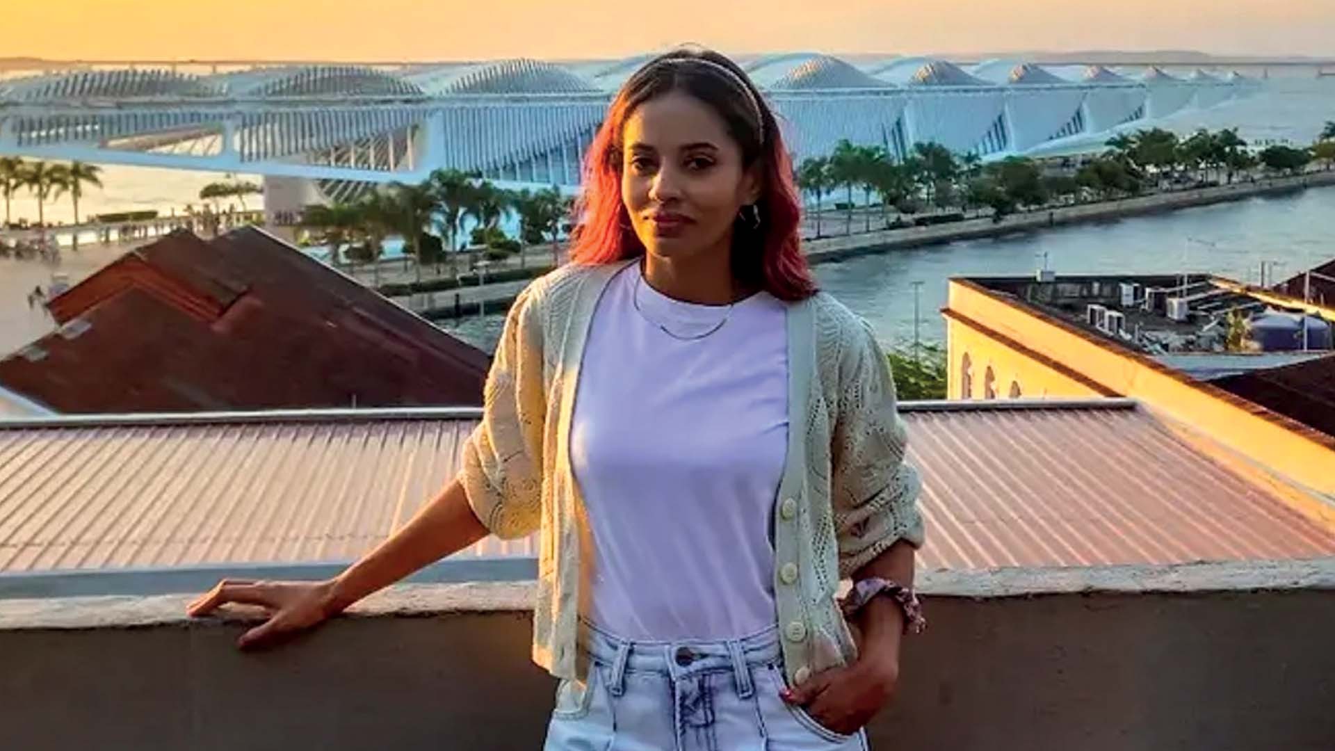 Mulher jovem com cabelo ruivo e tiara, sorrindo levemente, vestindo camiseta branca e cardigã bege, com a mão esquerda apoiada em um muro. Ao fundo, telhados, um rio e uma estrutura arquitetônica moderna com telhado ondulado, sob um céu alaranjado de pôr do sol