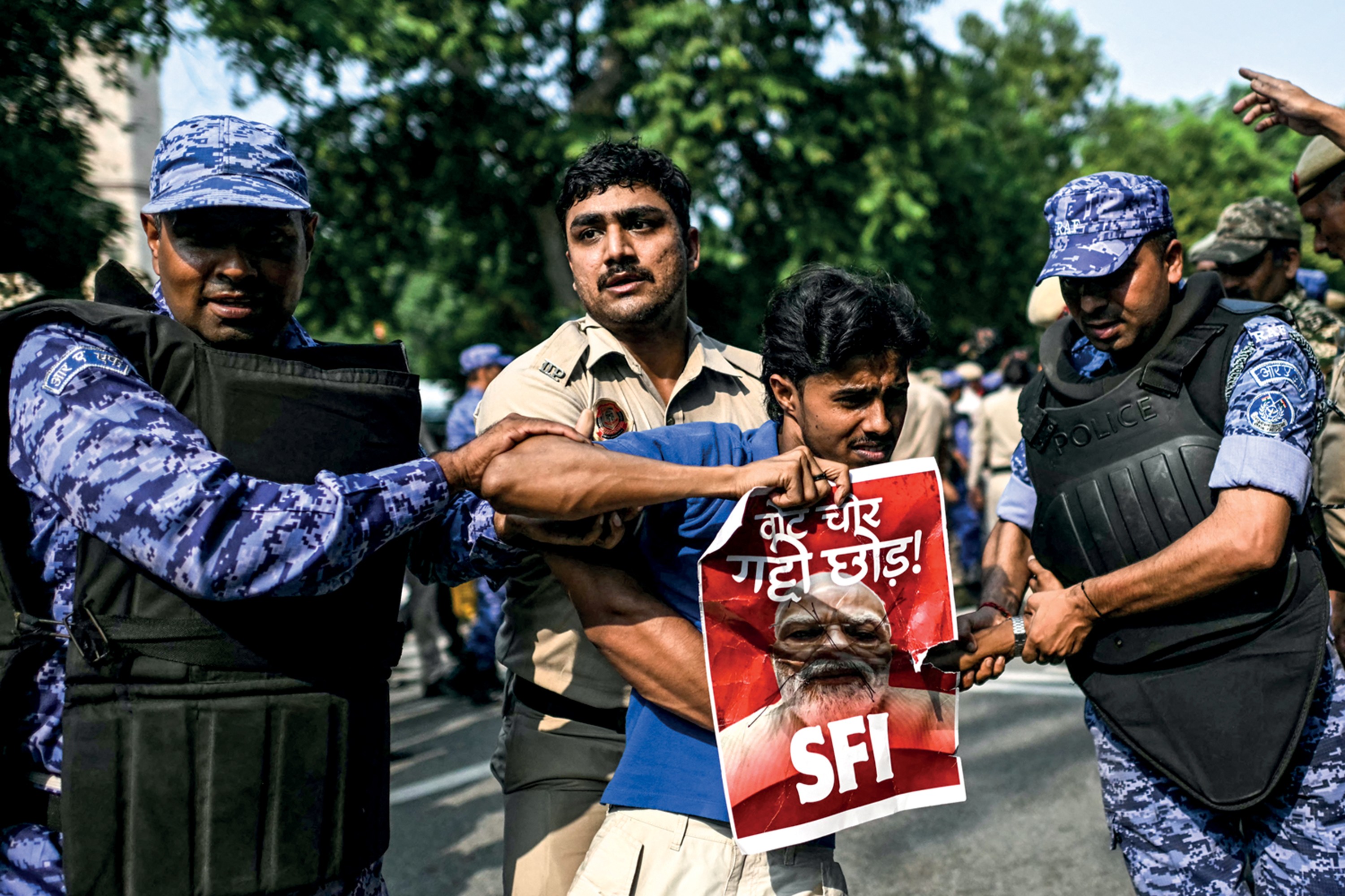 Três homens, dois policiais e um manifestante, em meio a uma multidão. O manifestante, no centro, segura um cartaz vermelho com a imagem de um homem barbudo e a sigla SFI. Um policial de farda camuflada azul e colete preto segura o braço do manifestante, enquanto outro policial de camisa bege o segura por trás.