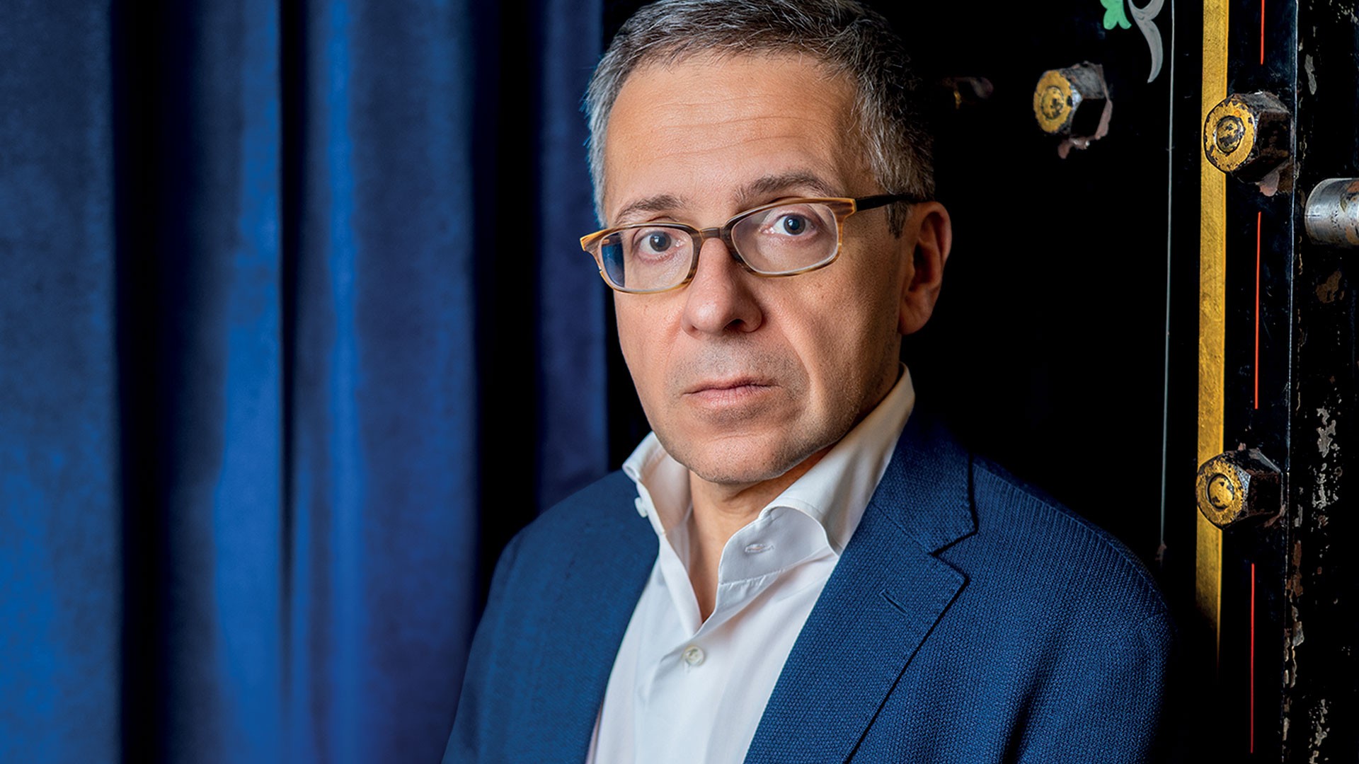 Homem de meia-idade, cabelos grisalhos e óculos, vestindo blazer azul e camisa branca, com expressão séria, posa contra cortina azul escura e uma porta de cofre antiga com detalhes dourados
