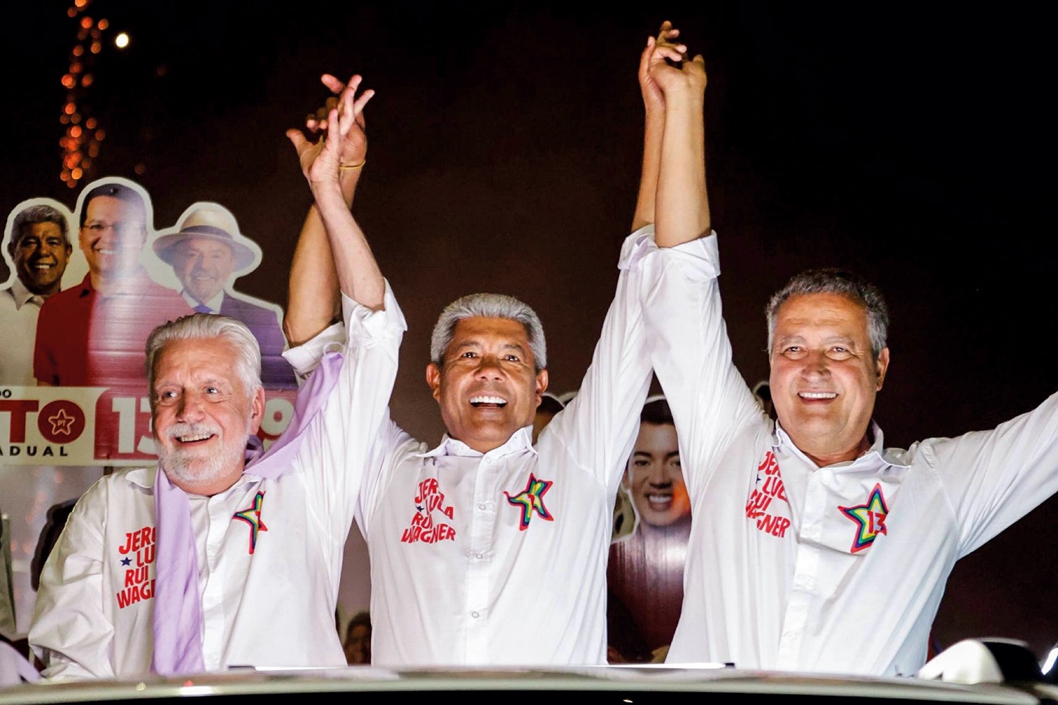 Três homens sorridentes, de camisas brancas, braços erguidos e mãos dadas, celebram em um comício noturno. O homem do centro, de cabelo grisalho, tem um broche de estrela colorida. O da esquerda, de barba branca, usa um lenço lilás. O da direita, de cabelo escuro, tem um broche de estrela vermelha. Ao fundo, um banner com rostos de outros políticos e o número 13