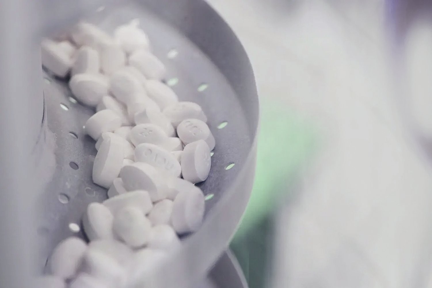 Comprimidos brancos e ovais, alguns com letras gravadas, sendo processados em uma máquina industrial metálica com furos, em ambiente de fábrica