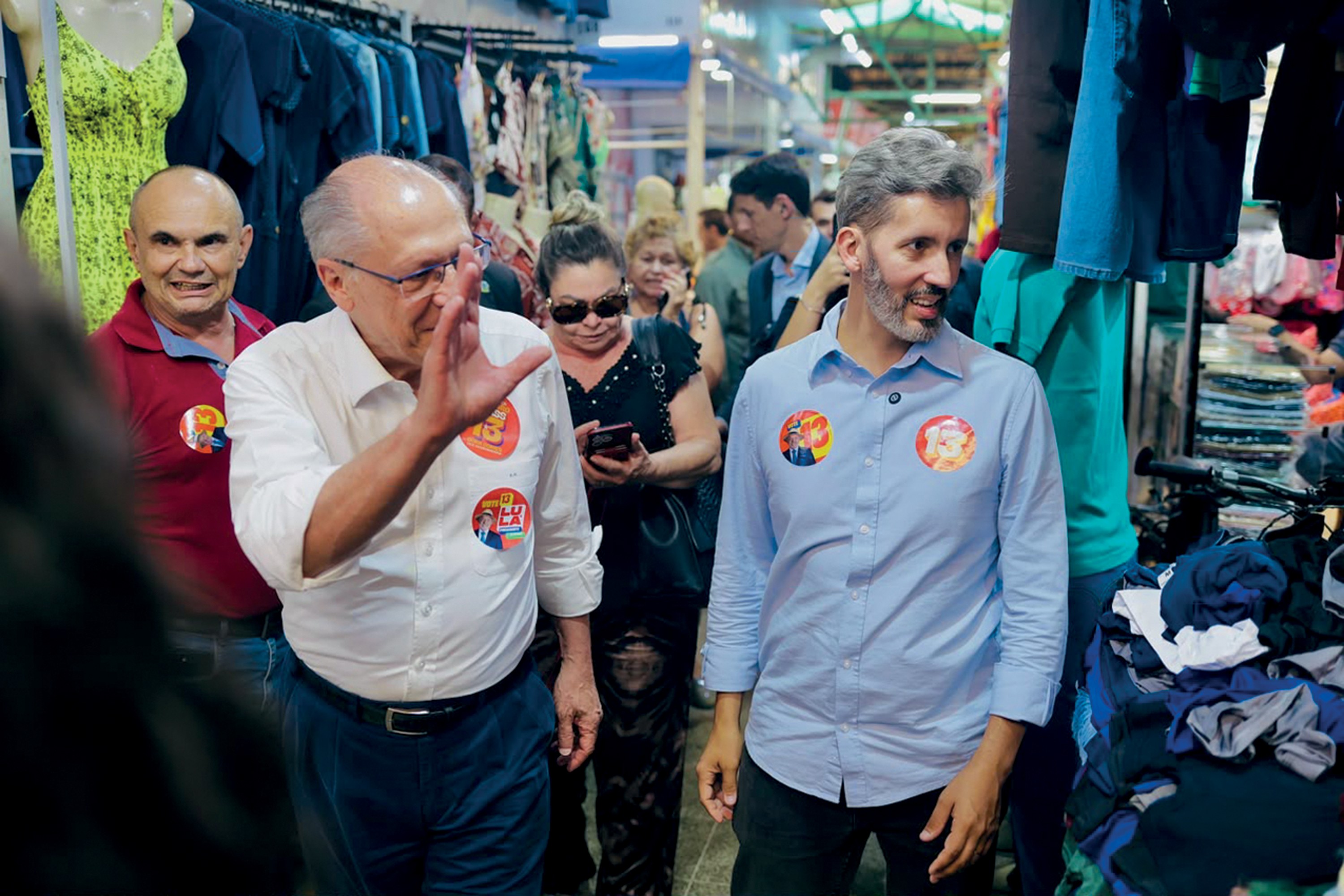 Homens em campanha política em um mercado popular. Um homem idoso de camisa branca acena, e um homem mais jovem de camisa azul sorri, ambos com broches de campanha. Outras pessoas e roupas penduradas ao fundo