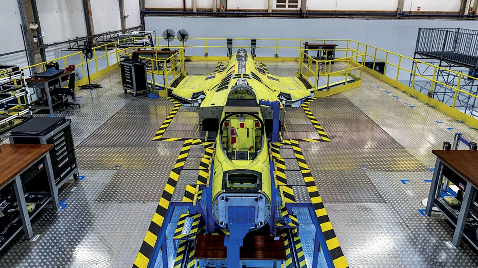 Um protótipo de avião de caça amarelo em uma linha de montagem industrial, com o cockpit aberto e estruturas de suporte azuis. O chão é de metal texturizado com faixas de segurança amarelas e pretas, e há grades amarelas ao redor da área de trabalho.