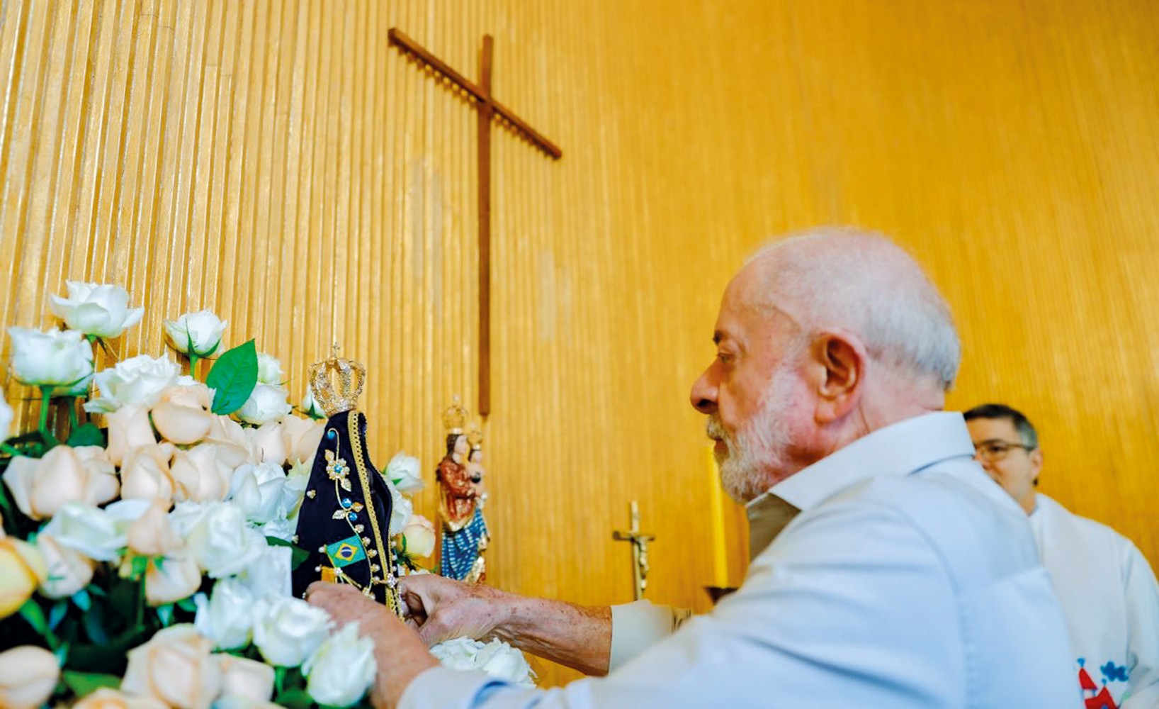 Lula, de perfil, com barba e cabelos brancos, veste camisa clara, tocando uma imagem de Nossa Senhora Aparecida com a bandeira do Brasil. Ao lado, um arranjo de rosas brancas e, ao fundo, uma cruz de madeira na parede amarela. Outro homem, de óculos, aparece desfocado atrás.
