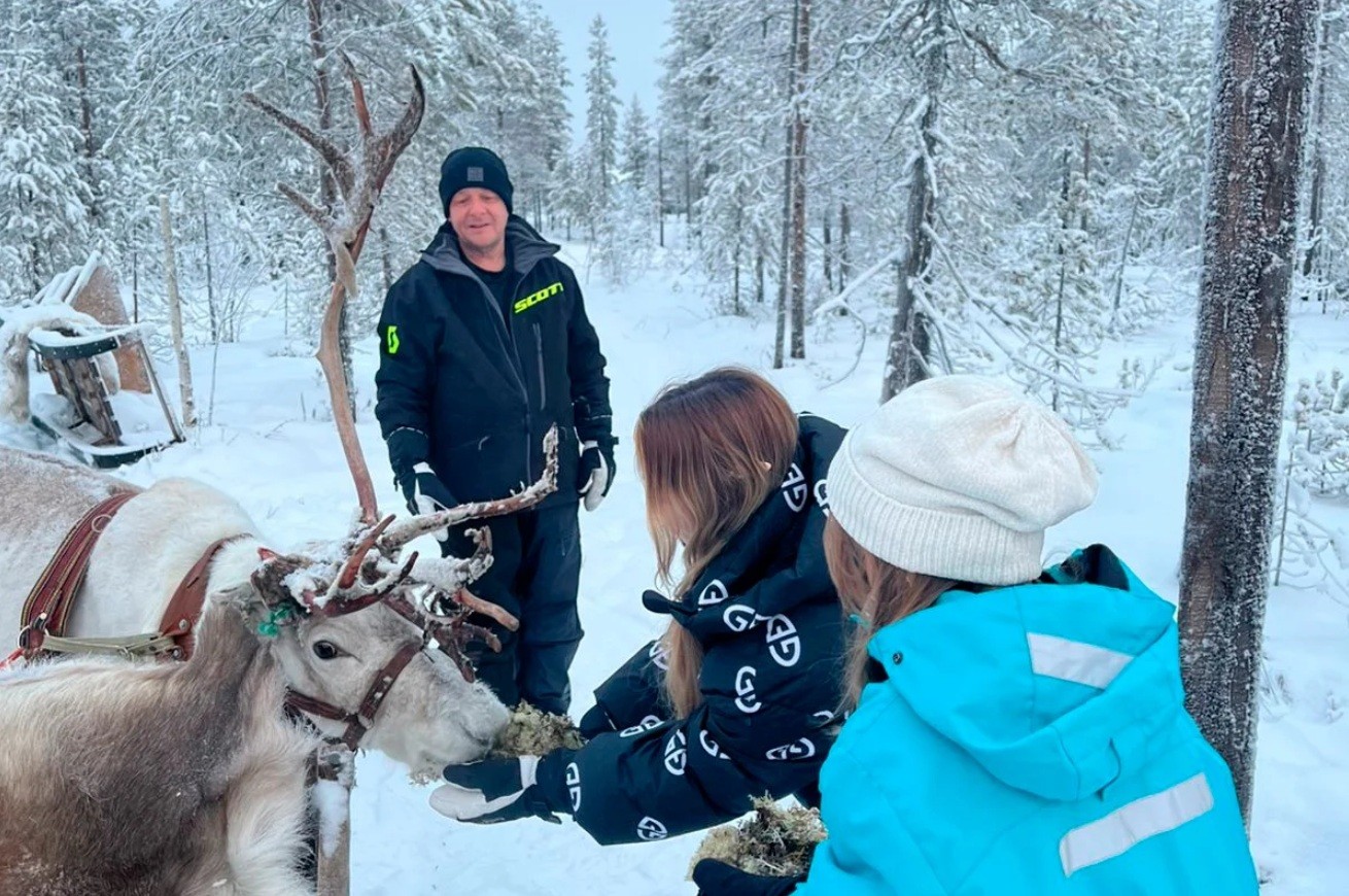 Um homem sorridente de jaqueta preta e gorro observa duas mulheres alimentando uma rena com musgo em uma floresta nevada. A rena está atrelada a um trenó, e as mulheres usam roupas de inverno, uma jaqueta preta com estampa e outra azul com gorro branco. O cenário é de inverno, com árvores cobertas de neve