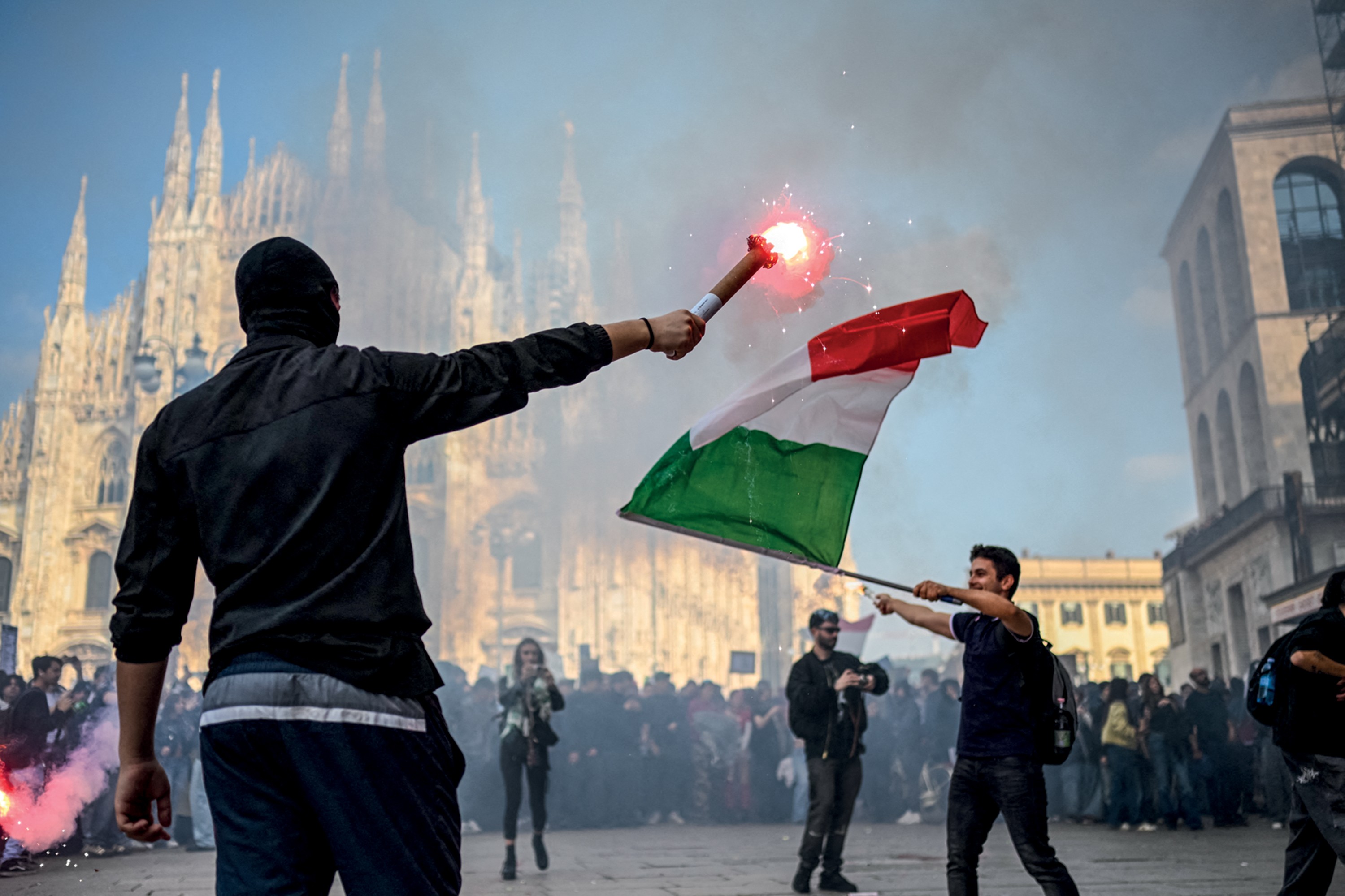Manifestantes em Milão, Itália, com a Catedral Duomo ao fundo. Um homem de costas, usando balaclava preta e jaqueta escura, segura um sinalizador vermelho aceso. Outro homem, à direita, agita uma bandeira italiana. Fumaça e uma multidão de pessoas são visíveis no cenário urbano.