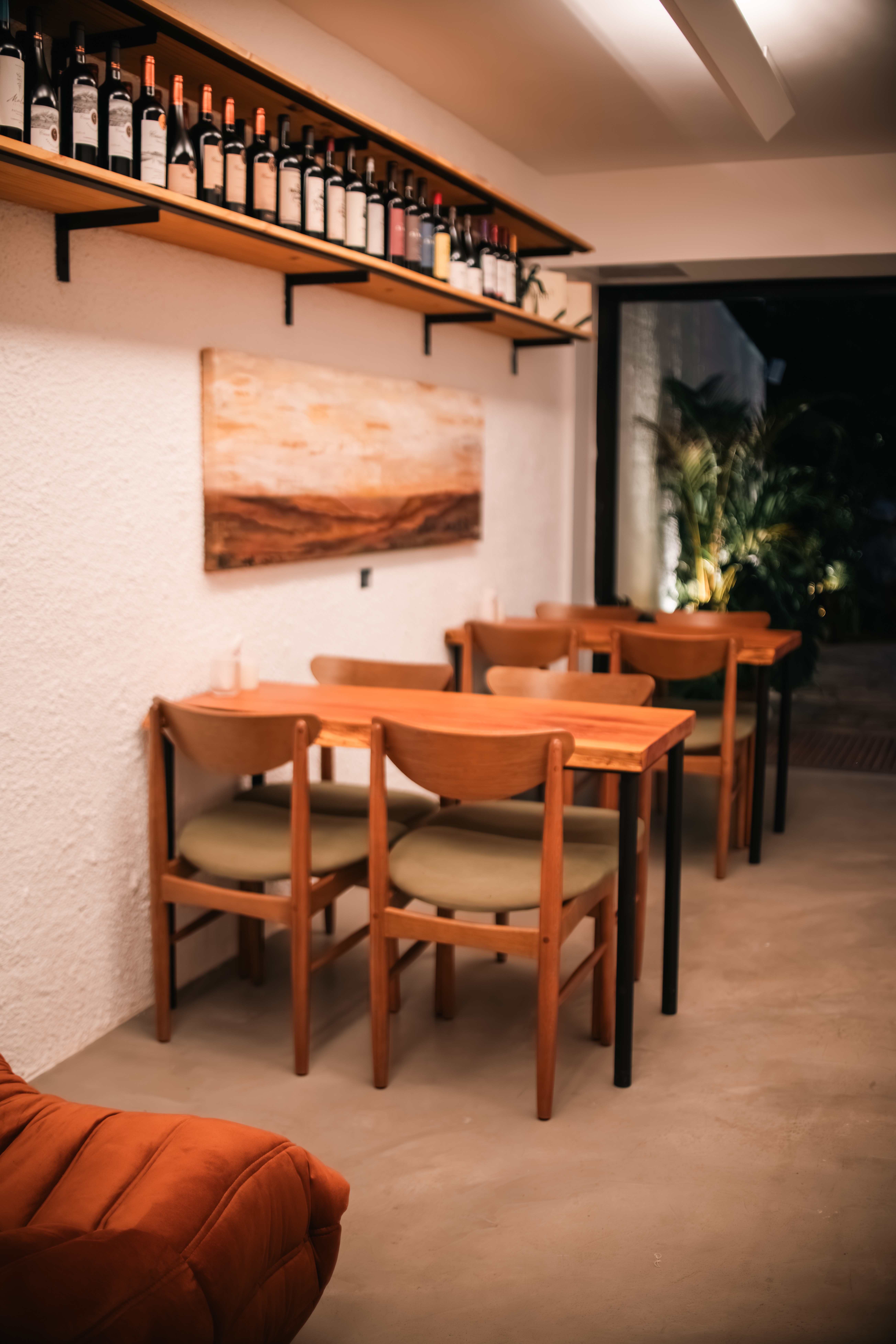 Interior de um restaurante com mesas e cadeiras de madeira clara e estofamento verde-oliva. Uma prateleira de madeira com garrafas de vinho est&aacute; acima de uma pintura abstrata em tons terrosos na parede branca. Um pufe laranja aparece no canto inferior esquerdo e plantas verdes s&atilde;o vis&iacute;veis atrav&eacute;s de uma porta escura ao fundo